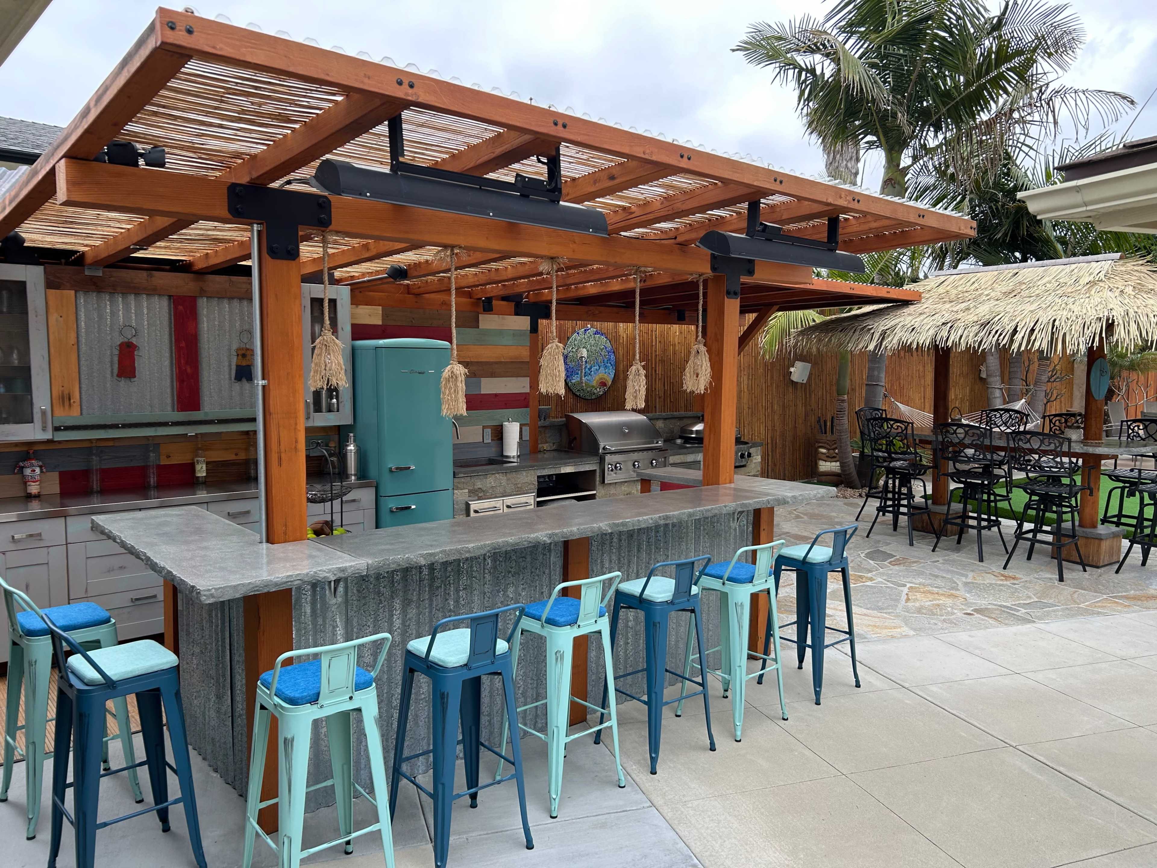 An outdoor kitchen and bar area featuring a stainless steel grill, a turquoise refrigerator, and several blue and teal stools arranged around a gray stone countertop, surrounded by tropical landscaping.