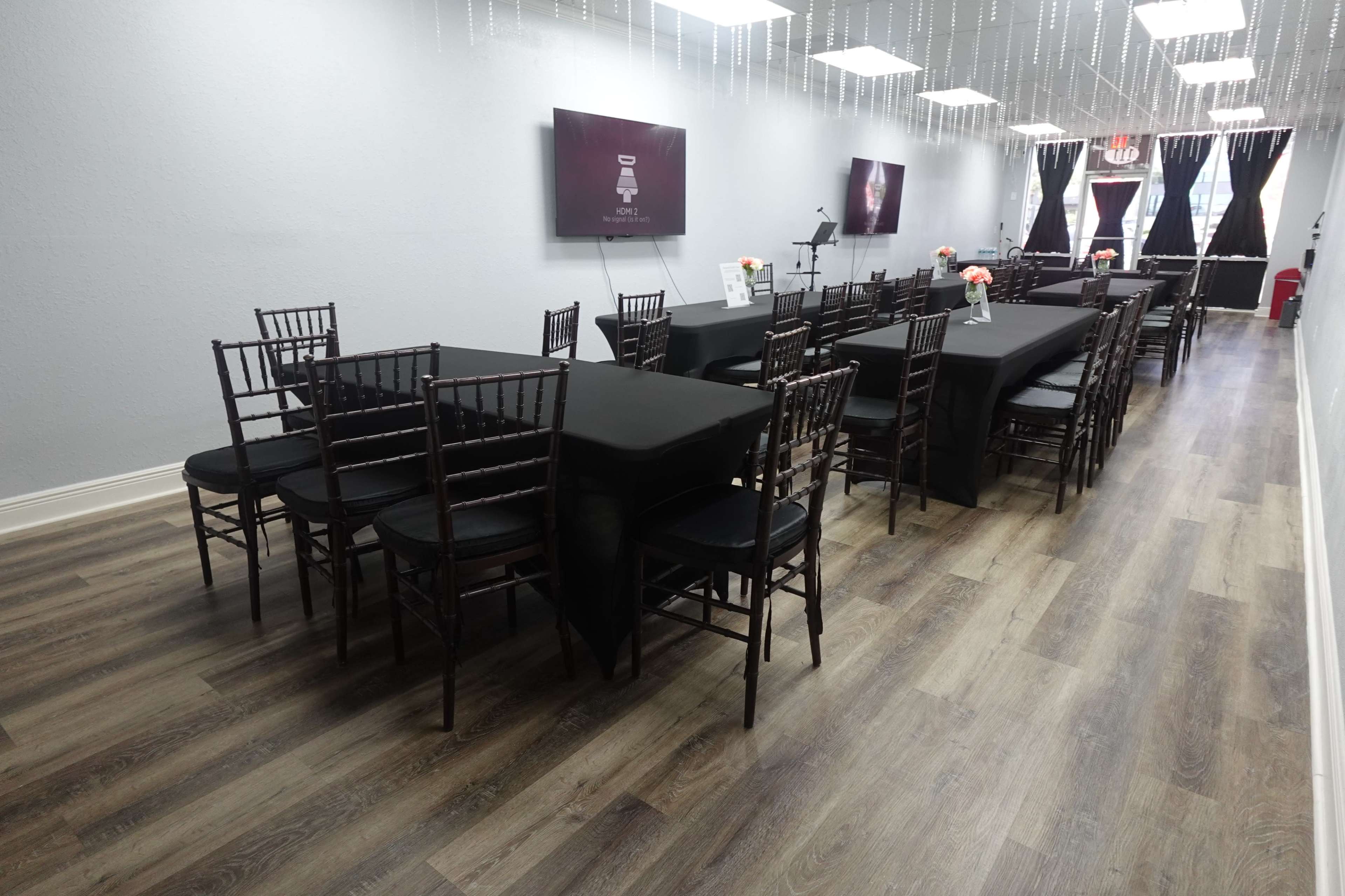 A spacious event room with black tables covered in tablecloths, surrounded by dark chiavari chairs, and decorated with floral centerpieces.