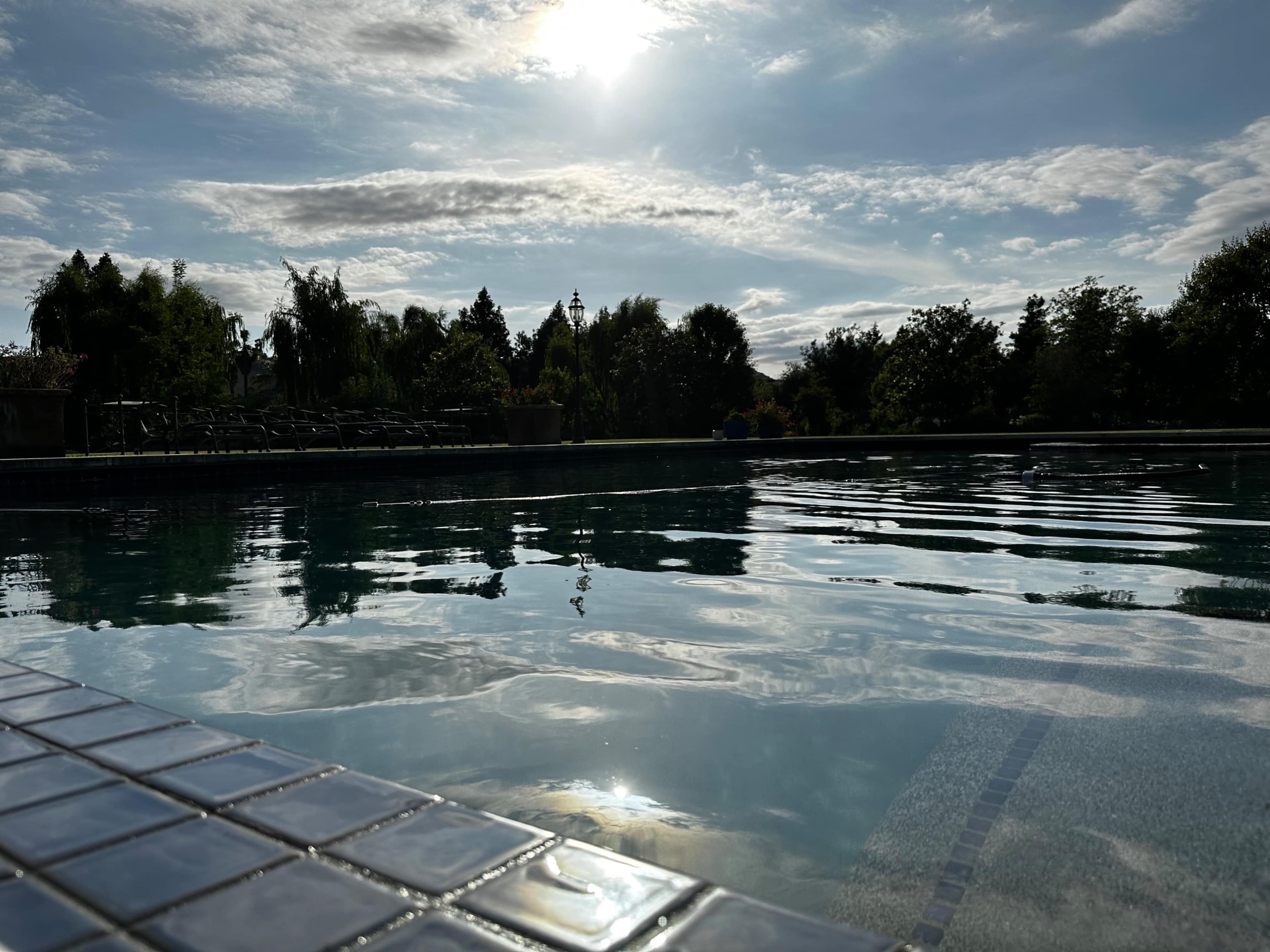A clear swimming pool with a reflection of clouds and the sun, surrounded by greenery and lounge chairs in the background.
