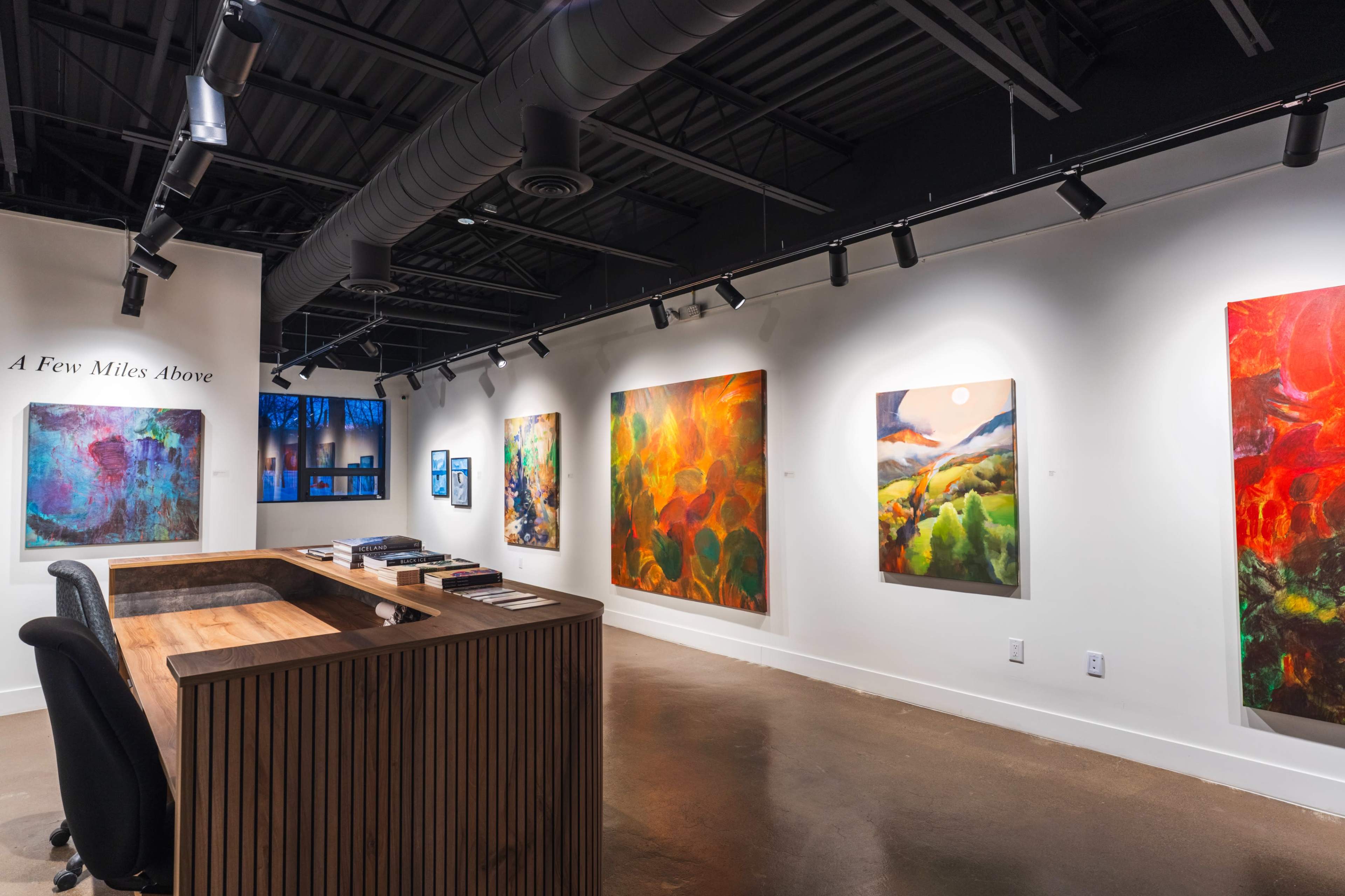 The image shows an art gallery interior featuring vibrant paintings on the walls, with a wooden reception desk in the foreground.