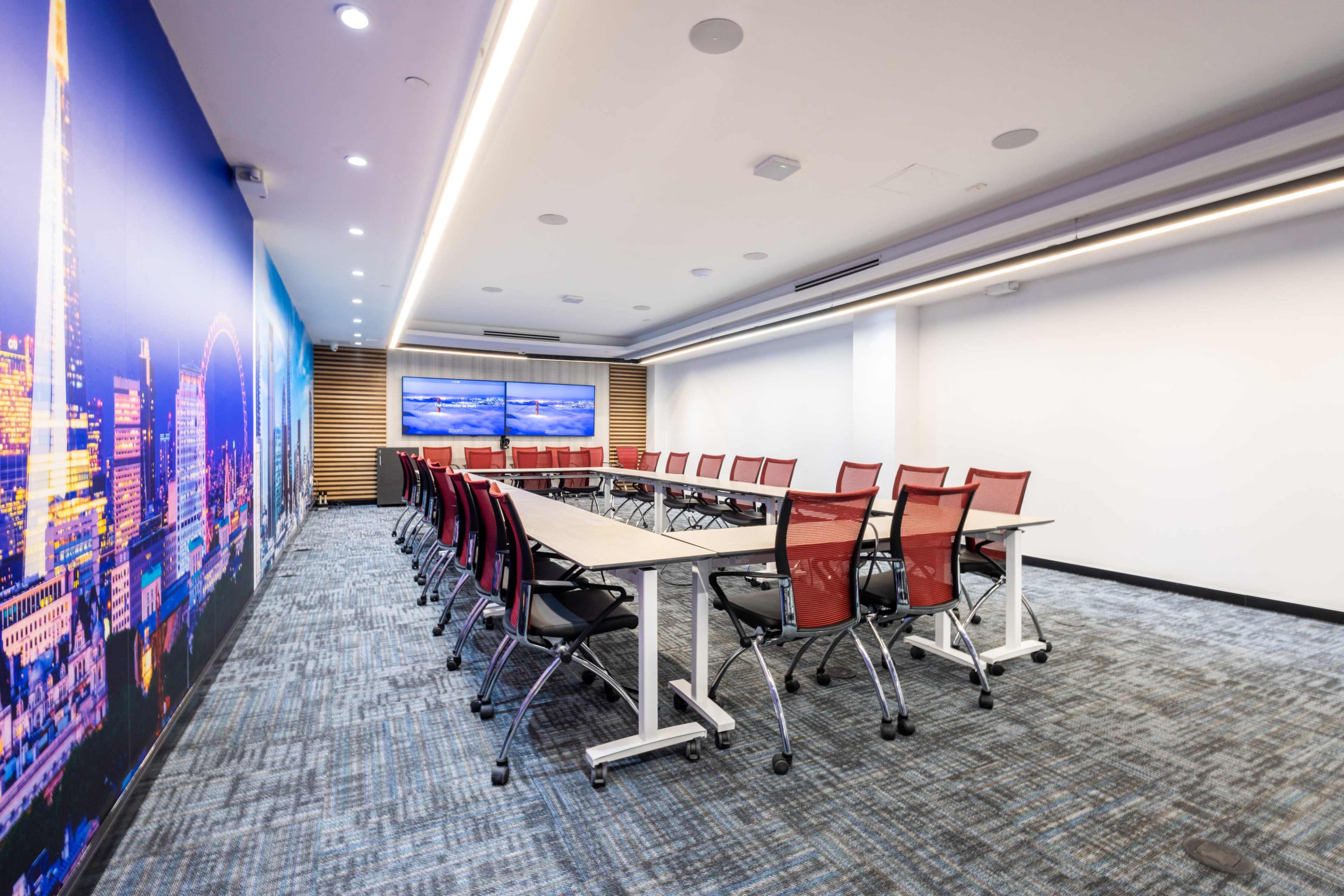 A modern conference room with a long table surrounded by red chairs, featuring large screens on the wall and a cityscape mural.