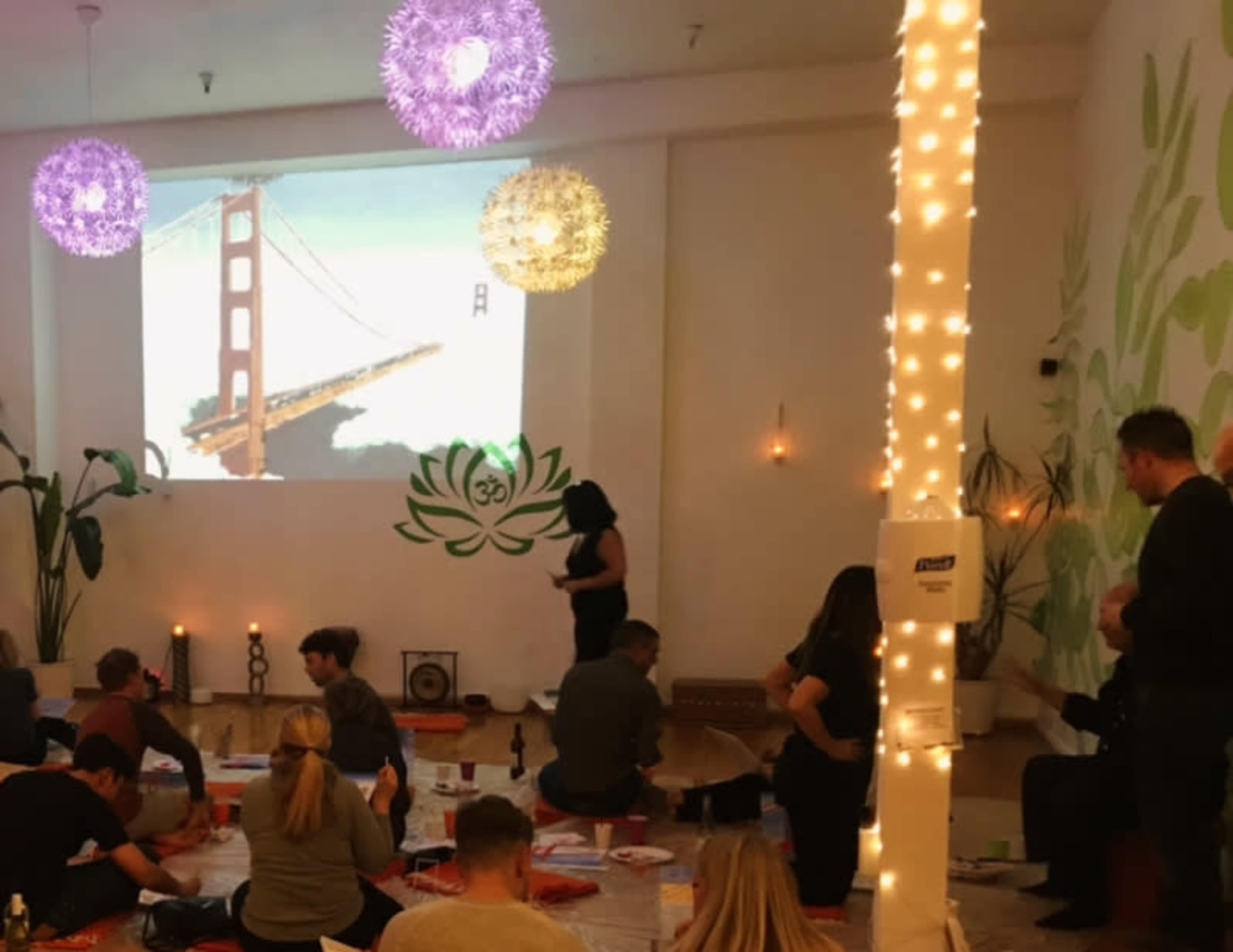 A group of people sits on the floor of a room with a large screen displaying an image of the Golden Gate Bridge, decorated with hanging lights and plants.