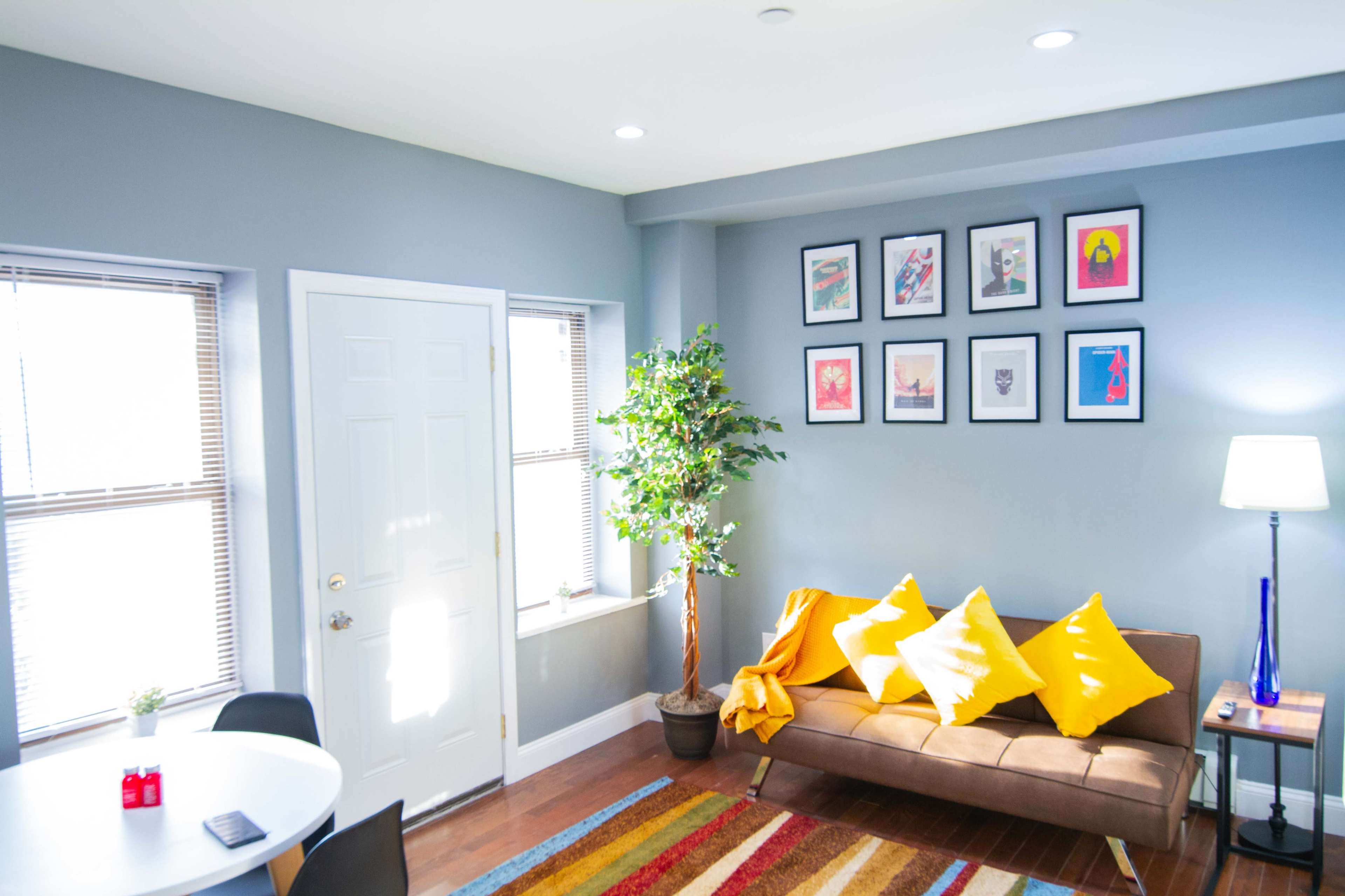 A cozy living room features a brown sofa with yellow cushions, a round dining table, colorful framed artwork on the walls, and a potted plant by the window.