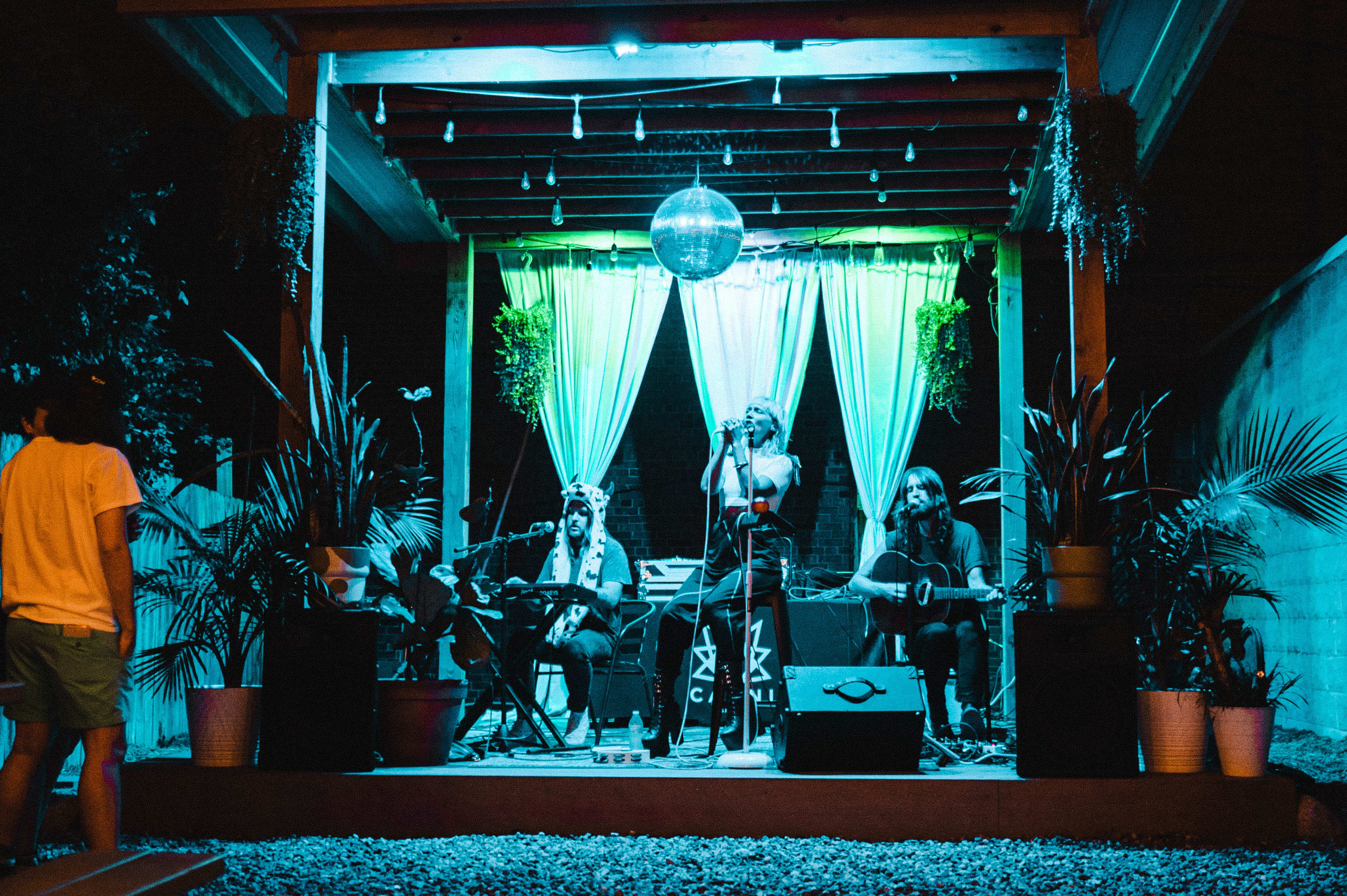 A live music performance takes place on a stage adorned with plants and colorful lighting, featuring musicians playing instruments under a disco ball.