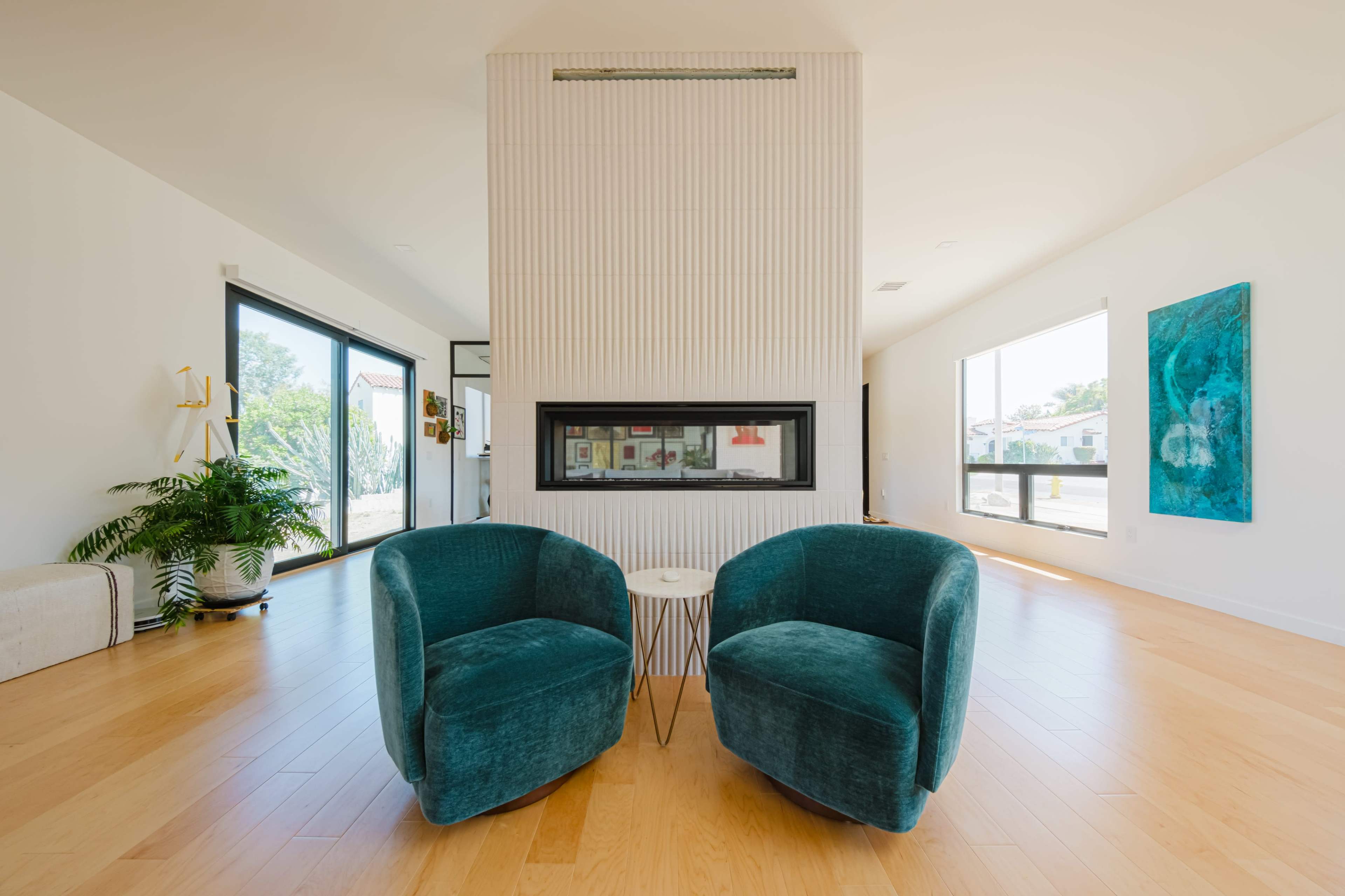 The image shows a modern living room with two teal chairs facing a central fireplace, surrounded by large windows and minimalistic decor.