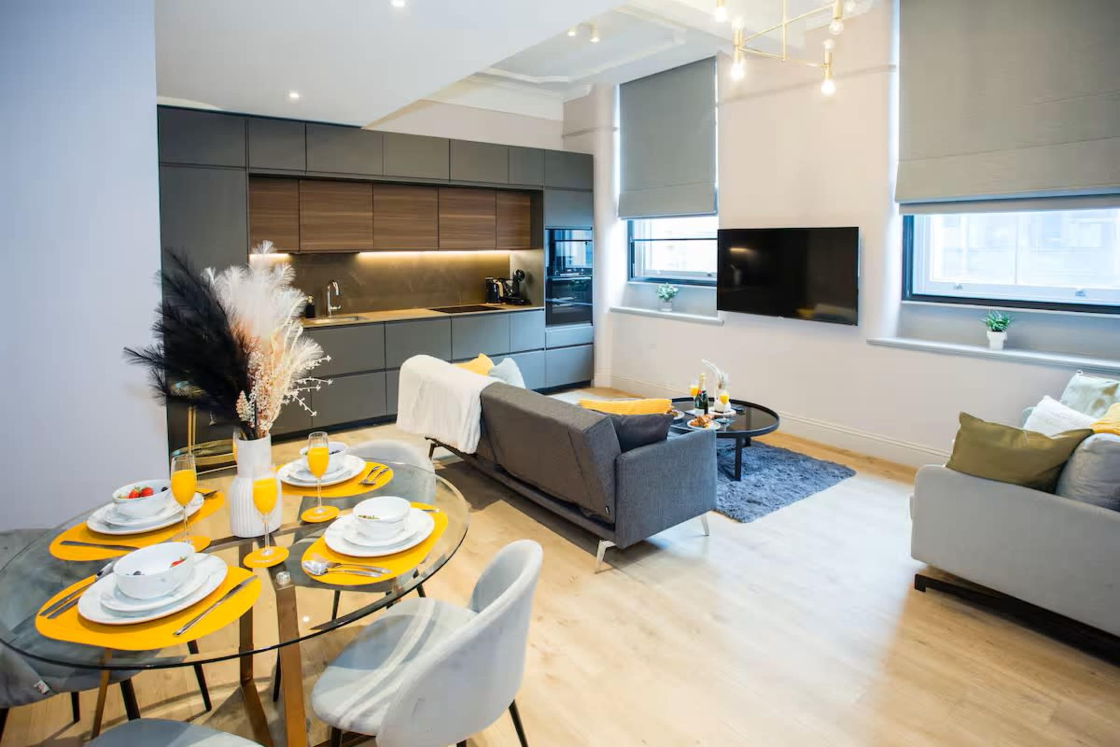 The image shows a modern open-plan living and dining area with a kitchen, featuring a round dining table set for breakfast, a cozy sitting area with a sofa and décor, and large windows allowing natural light.