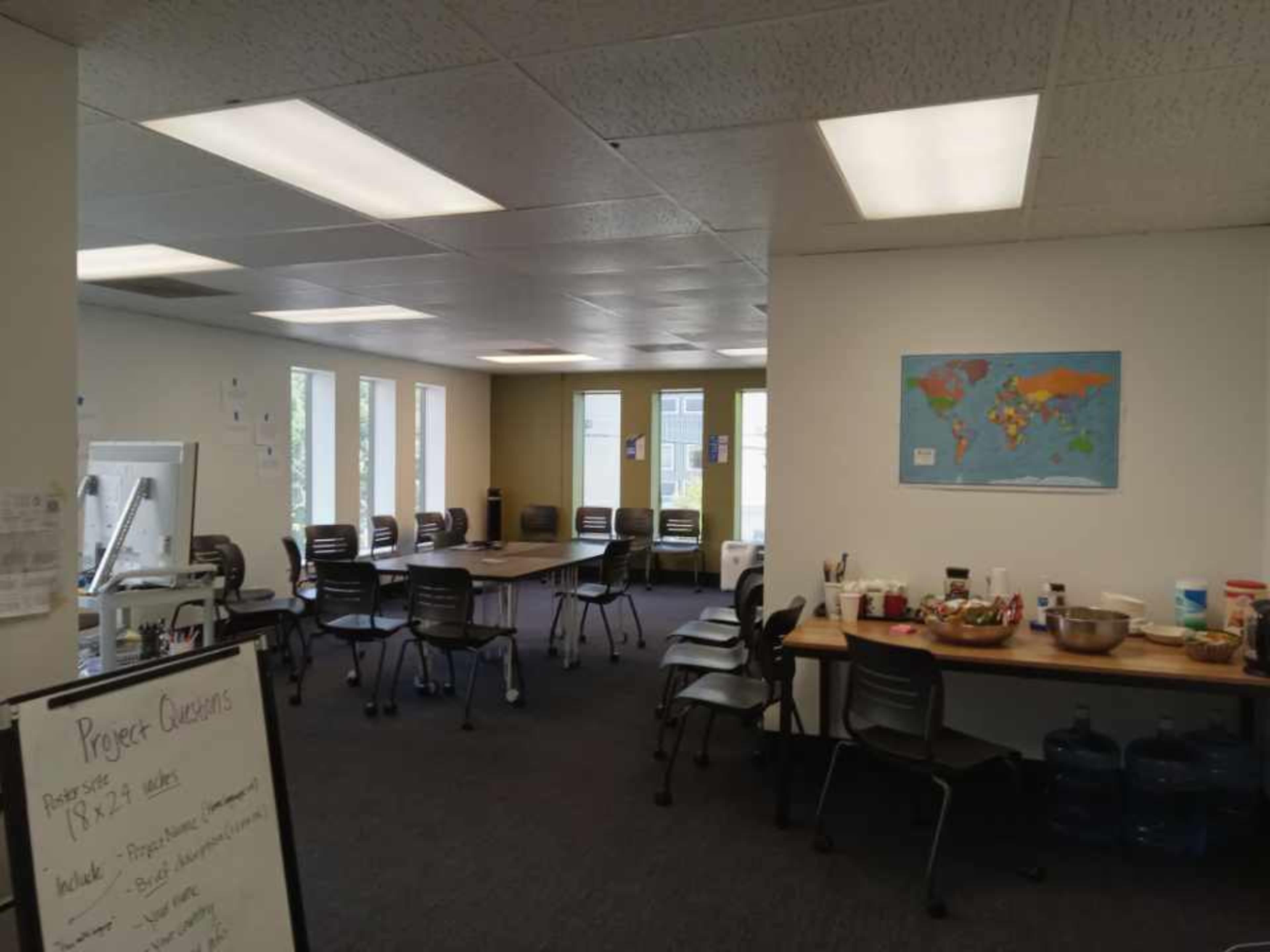 A spacious office area features multiple tables and chairs, large windows, and a world map displayed on the wall.