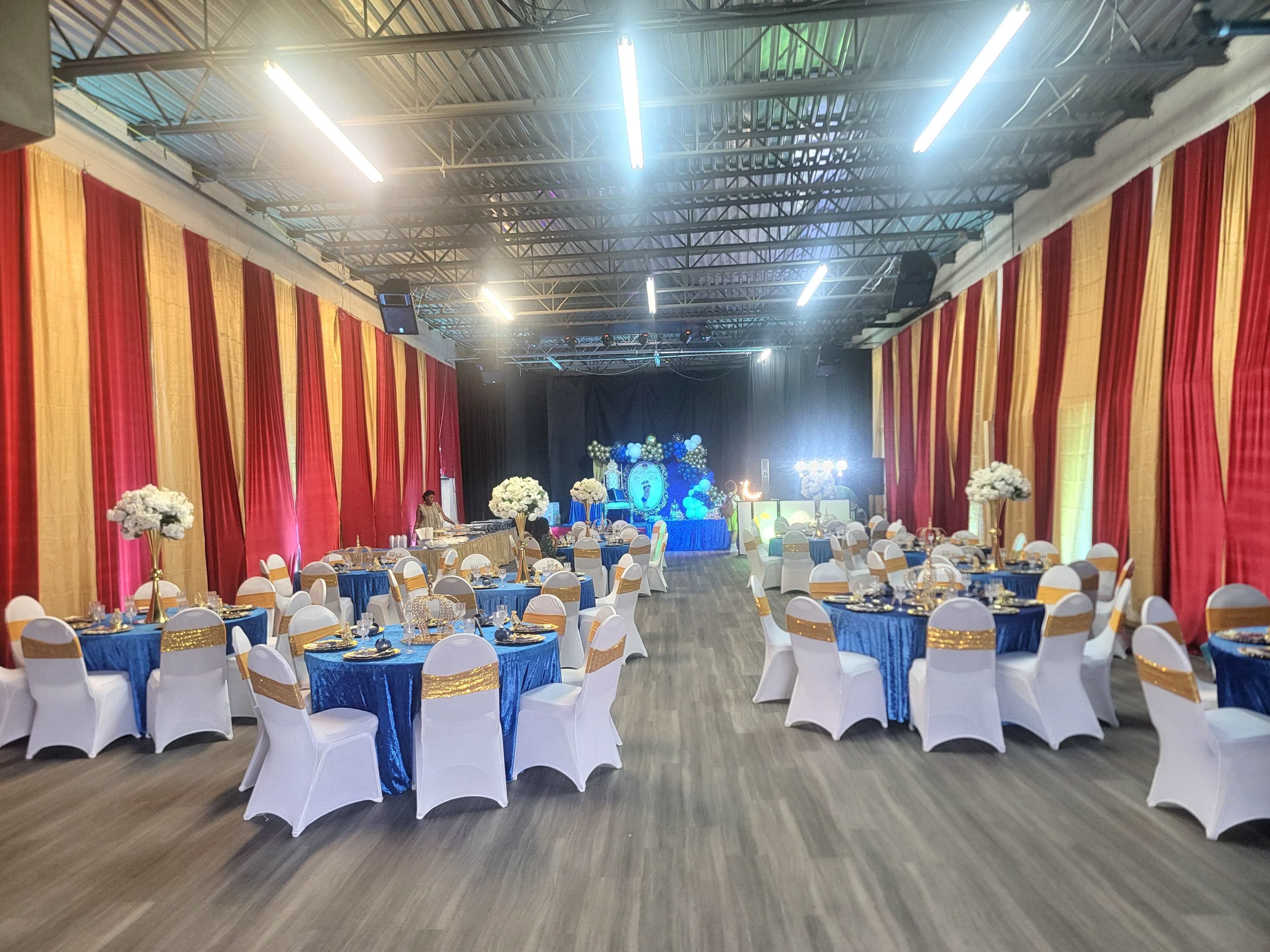 The image shows a banquet hall decorated with blue and gold tablecloths, white chairs, and draped red and gold fabric on the walls, set for an event.