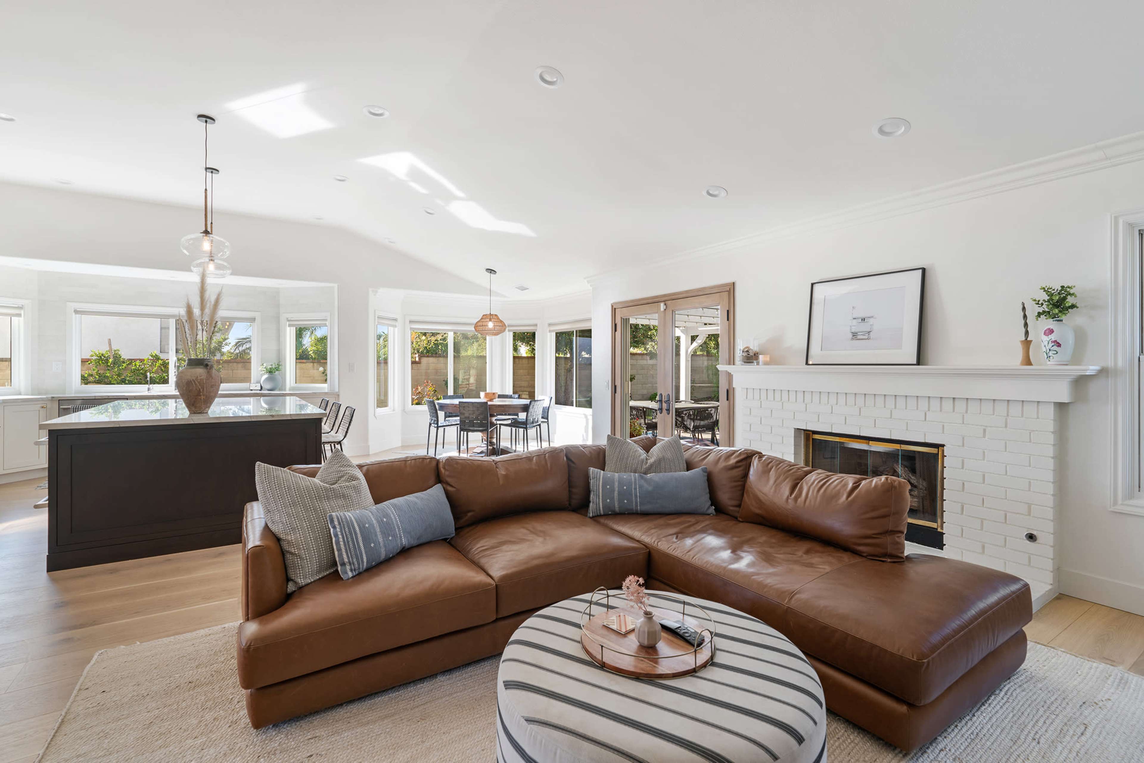 A spacious living room features a brown leather sectional sofa, a round coffee table, and large windows overlooking a well-lit dining area.