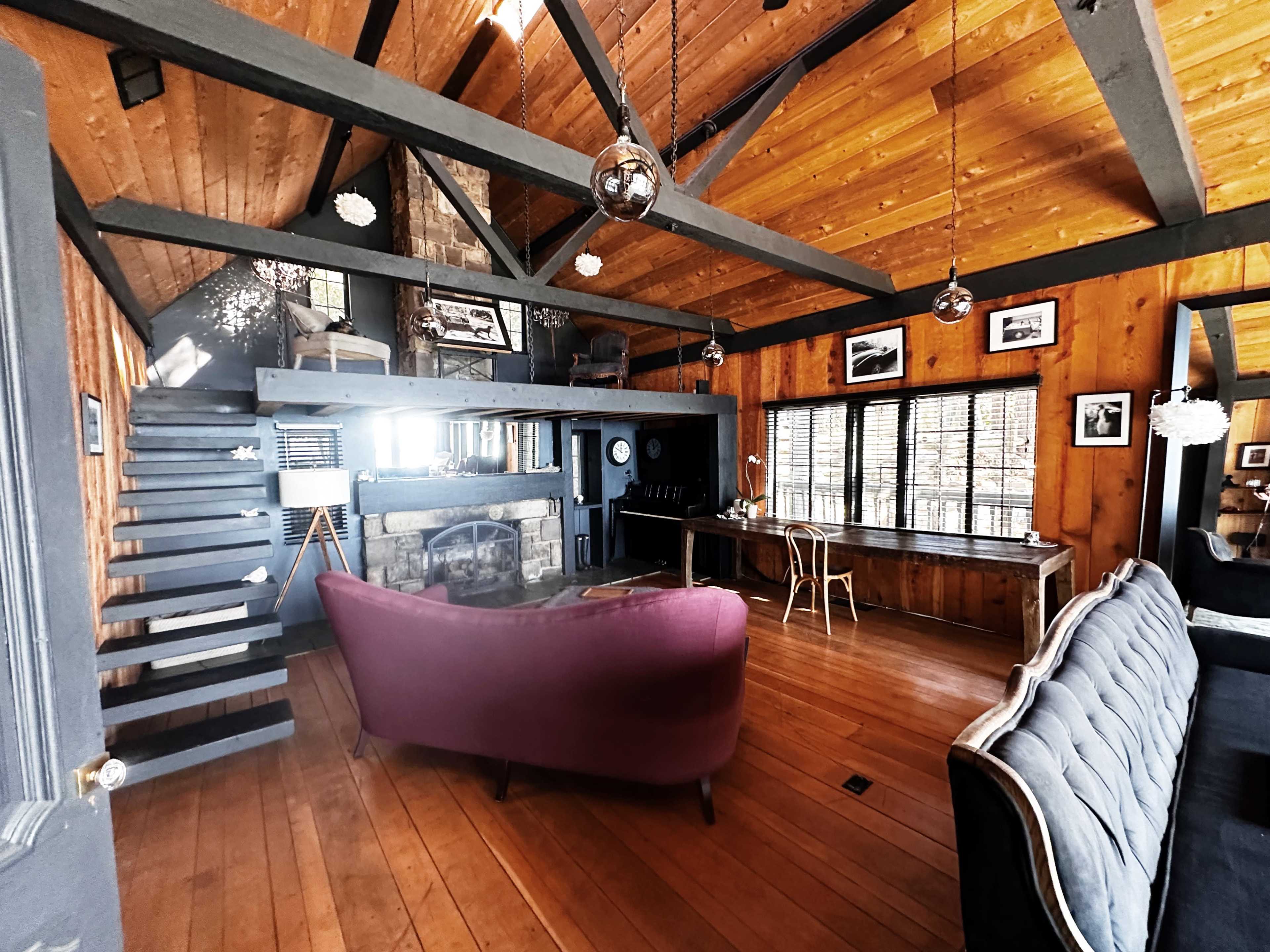 A spacious wooden interior of a cabin featuring a staircase, a purple sofa, and a large stone fireplace under a high ceiling with exposed beams.