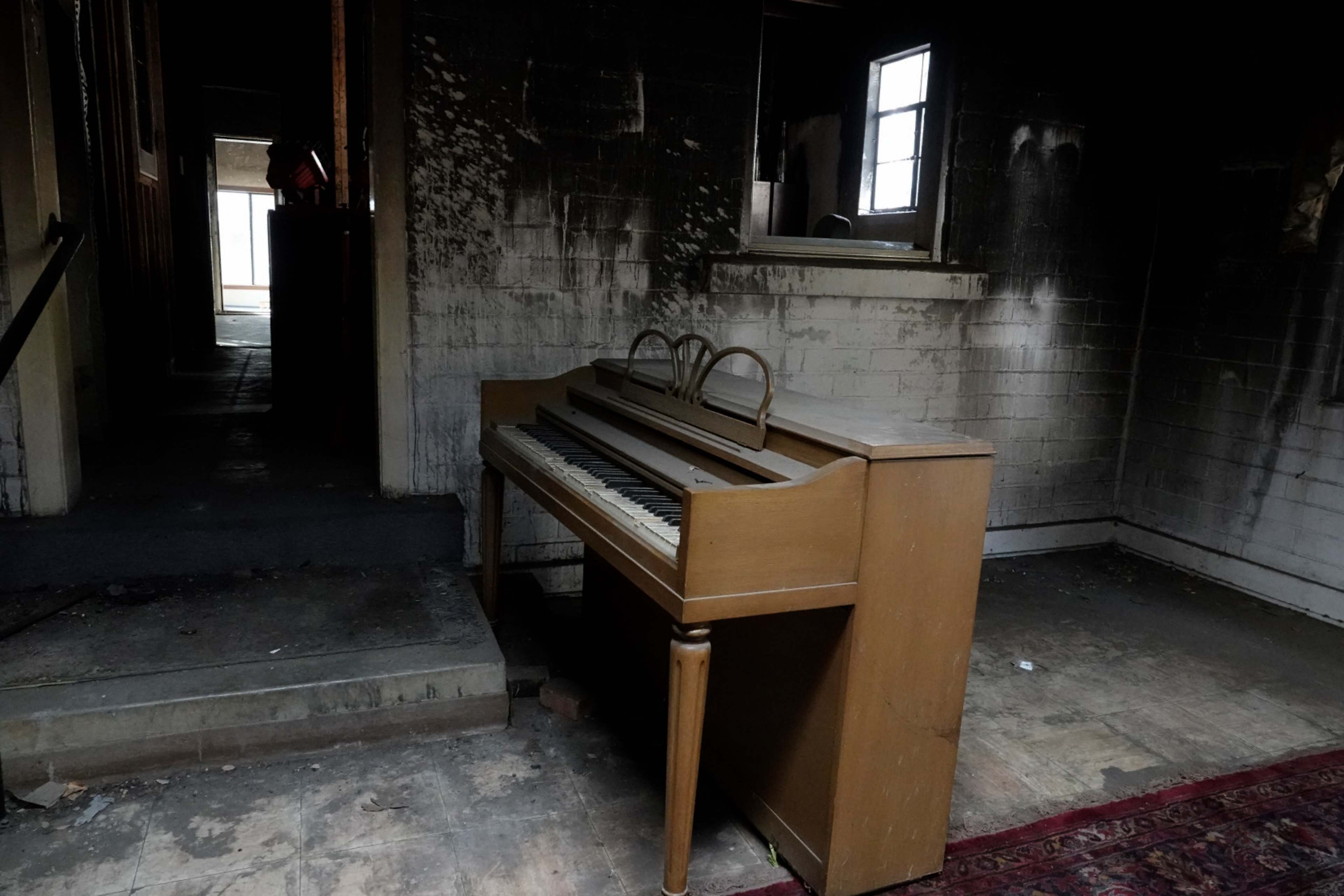 An old piano stands in a dimly lit, abandoned room with stained walls and scattered debris.