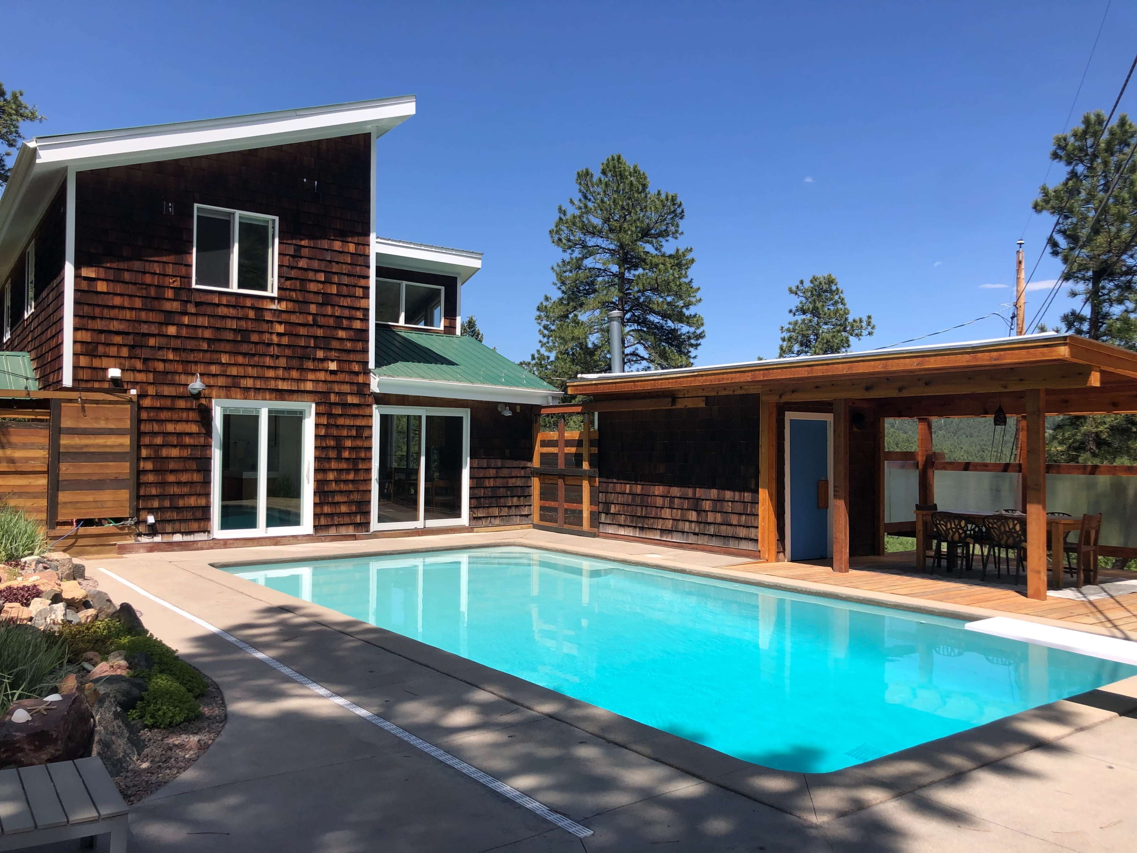 A modern home with a wooden exterior and a green roof is adjacent to a clear blue swimming pool and a shaded outdoor dining area.