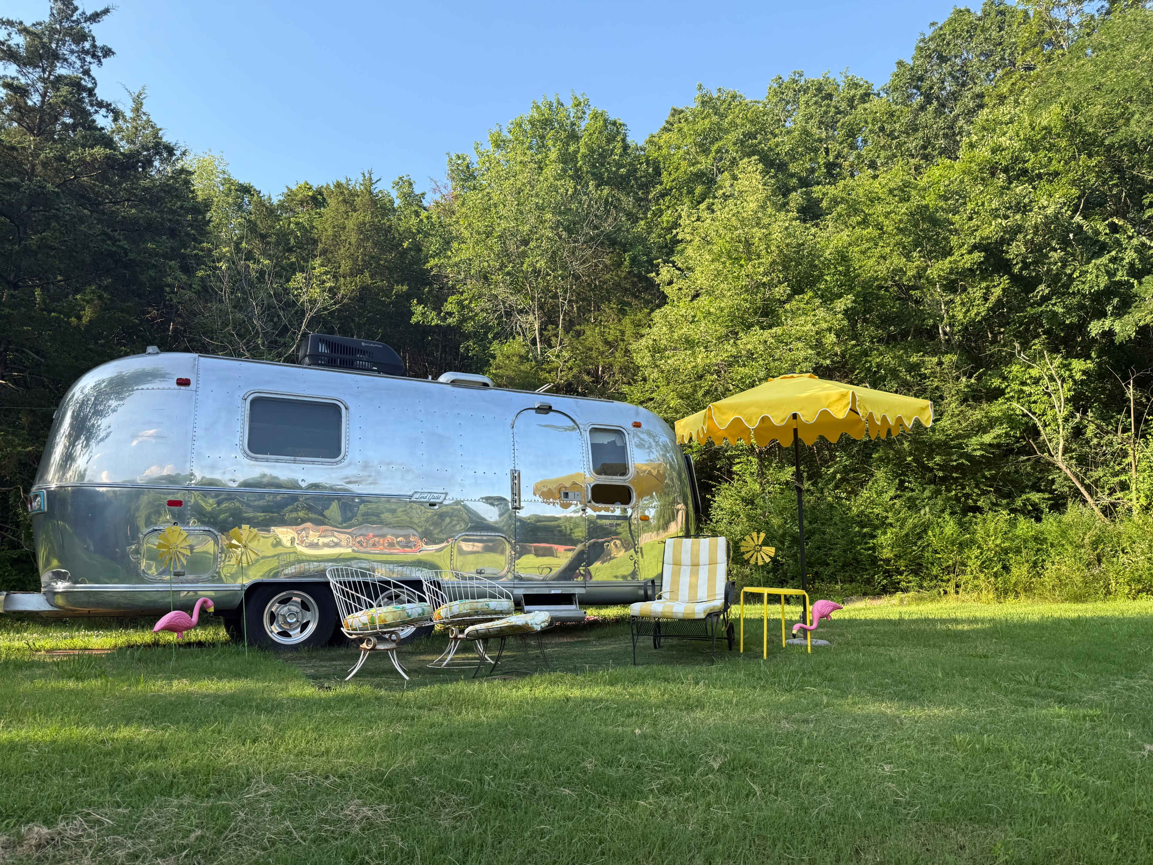A shiny Airstream trailer is parked on green grass beside a yellow patio umbrella, striped chairs, and pink flamingo lawn ornaments under a wooded area.