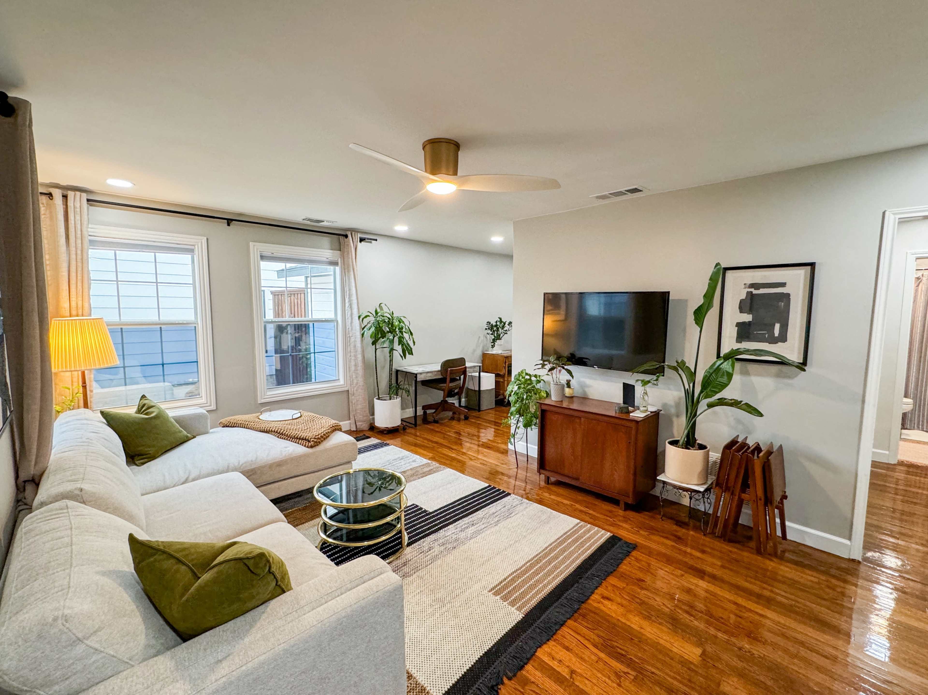 The living room features a light-colored sectional sofa, a wooden coffee table, a television mounted on the wall, and plants arranged around the space.