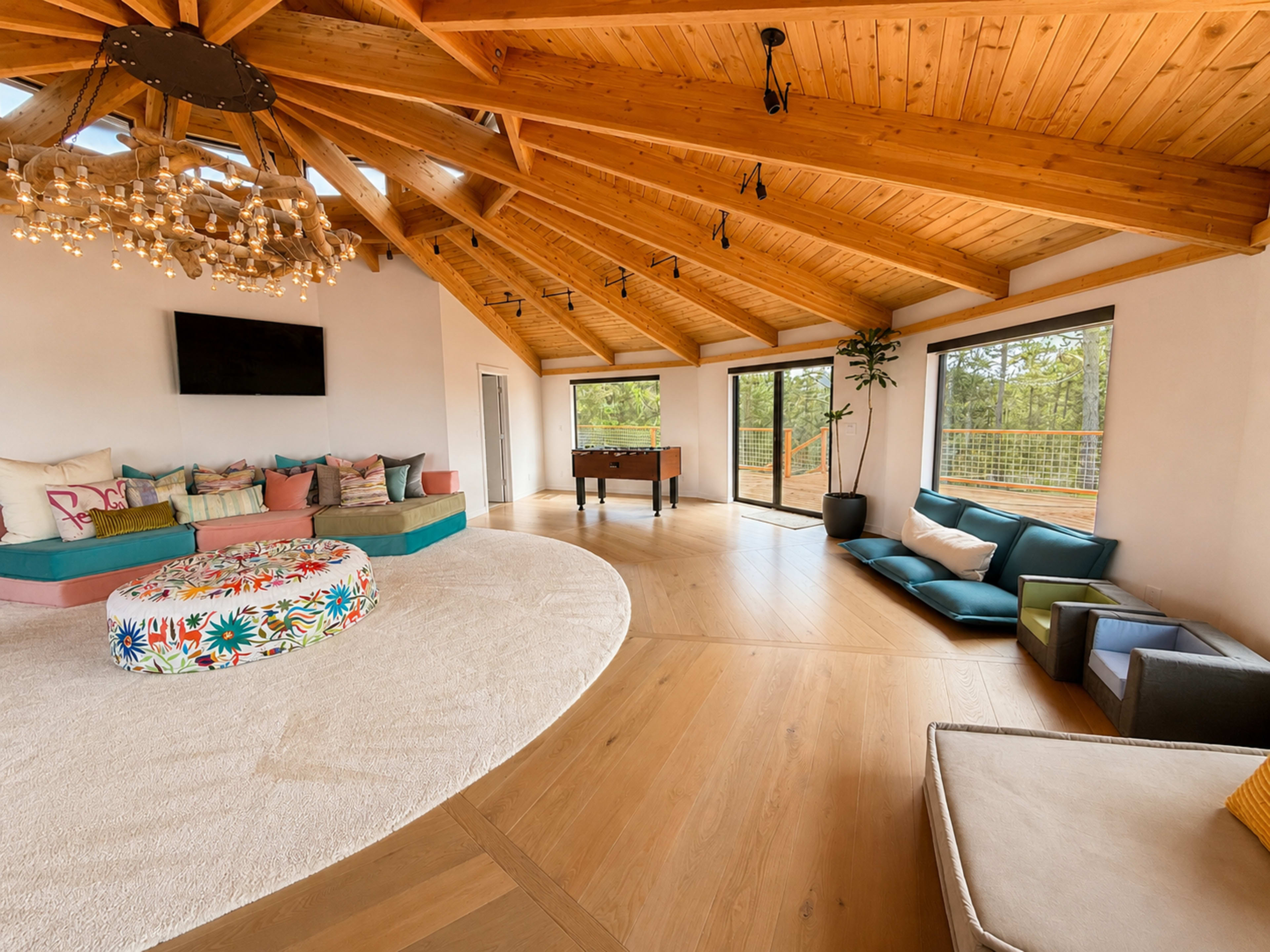 The image shows a spacious, modern living room with wooden ceilings, a circular area rug, colorful seating arrangements, a foosball table, and large windows that provide natural light.