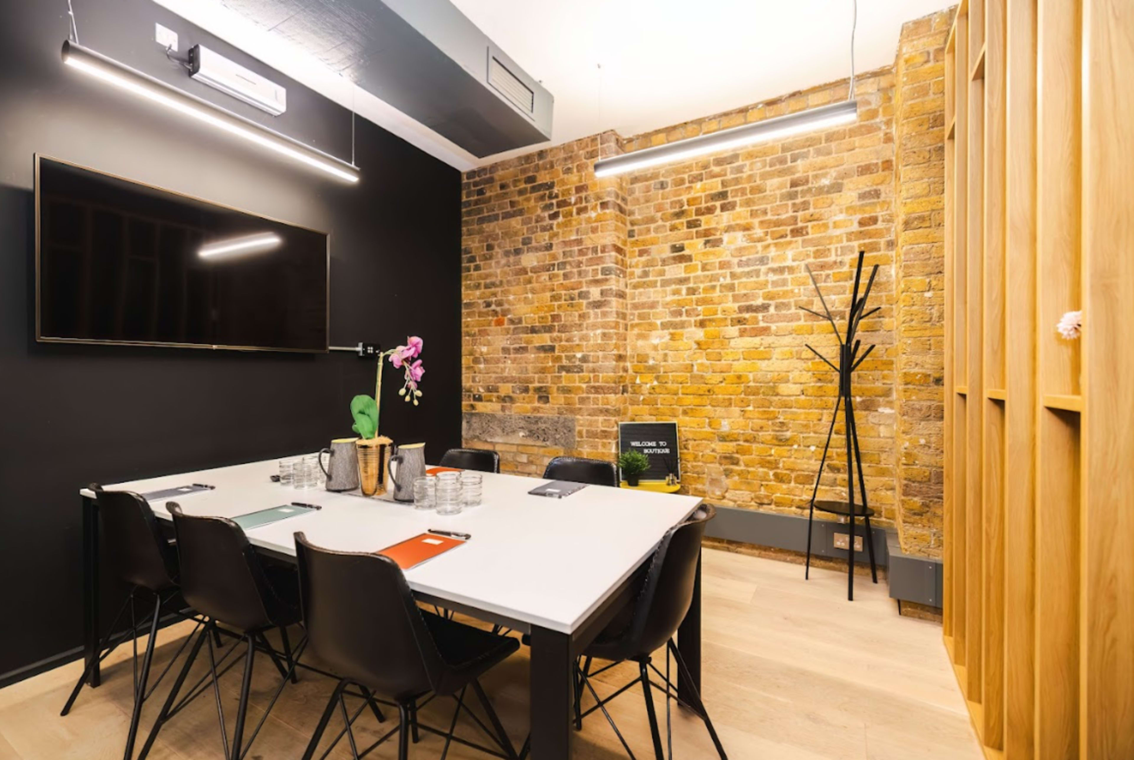 A conference room features a large table, black chairs, a wall-mounted TV, and exposed brick walls.