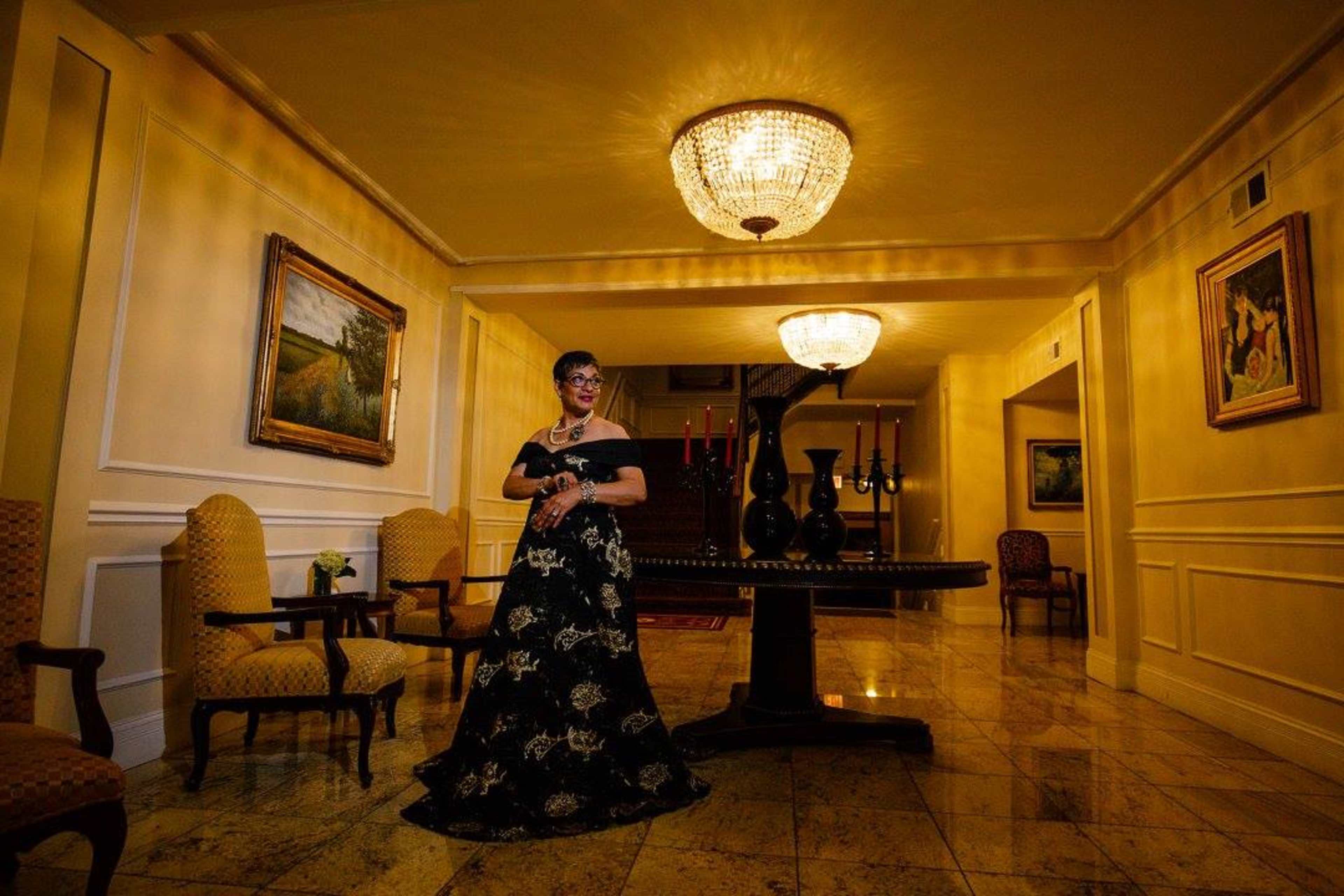 A woman in an elegant black dress stands beside a large table under a chandelier in a well-lit hallway adorned with paintings and chairs.