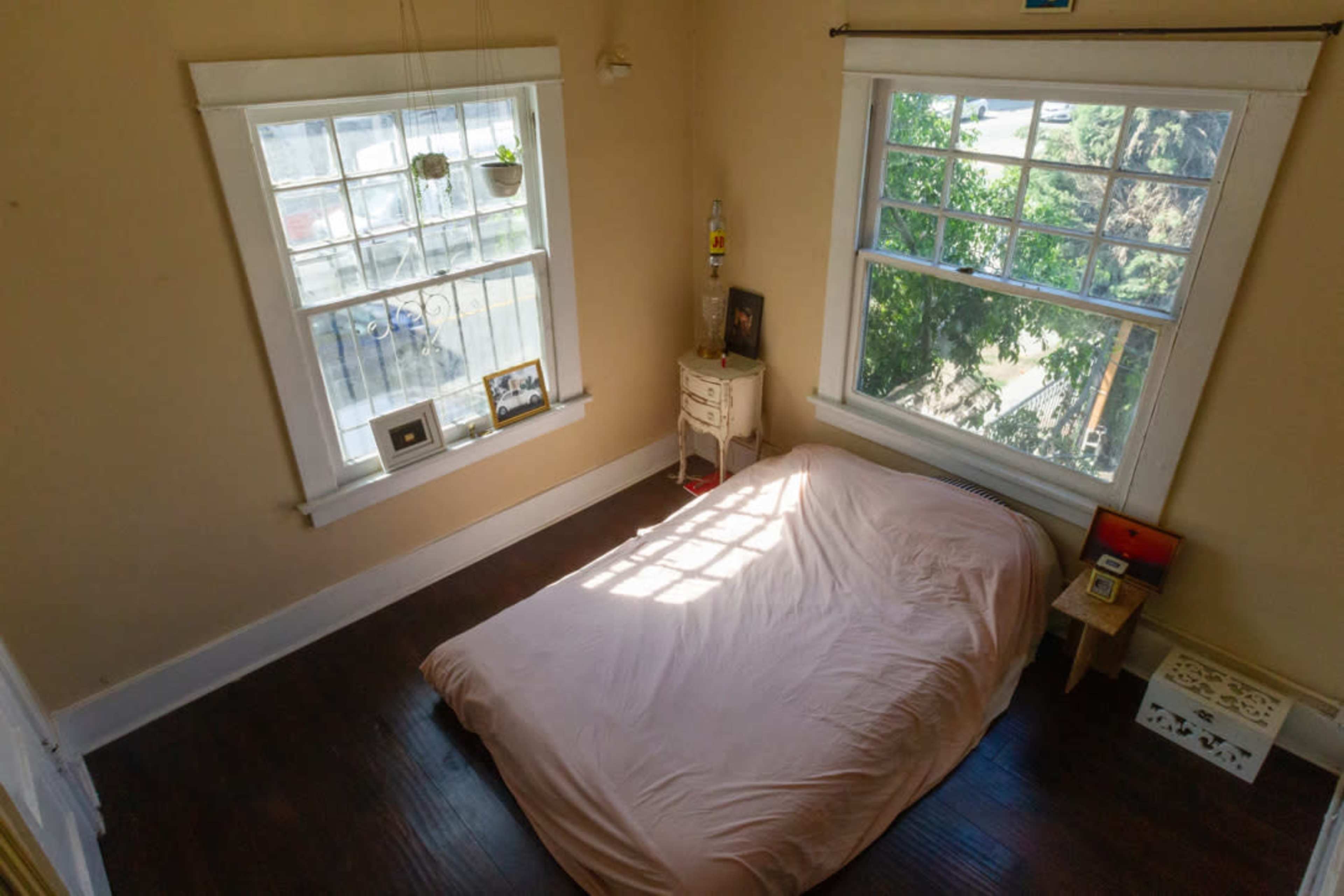 A simple bedroom with a bed covered in a pink sheet, two windows letting in light, and minimal furnishings including a small table and plants.