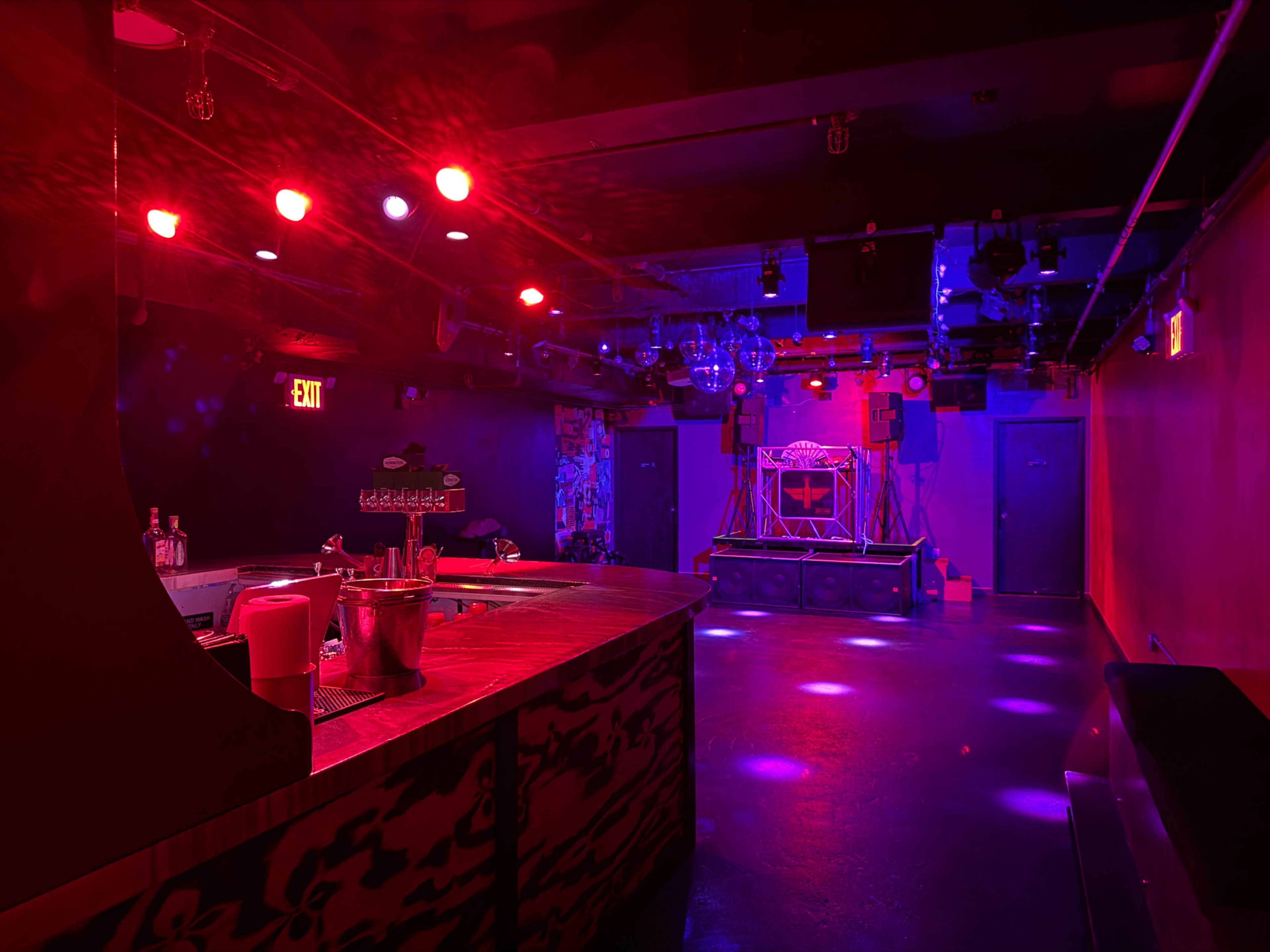 An empty bar area with a stage at the far end, illuminated by colorful lights in a dimly lit room.