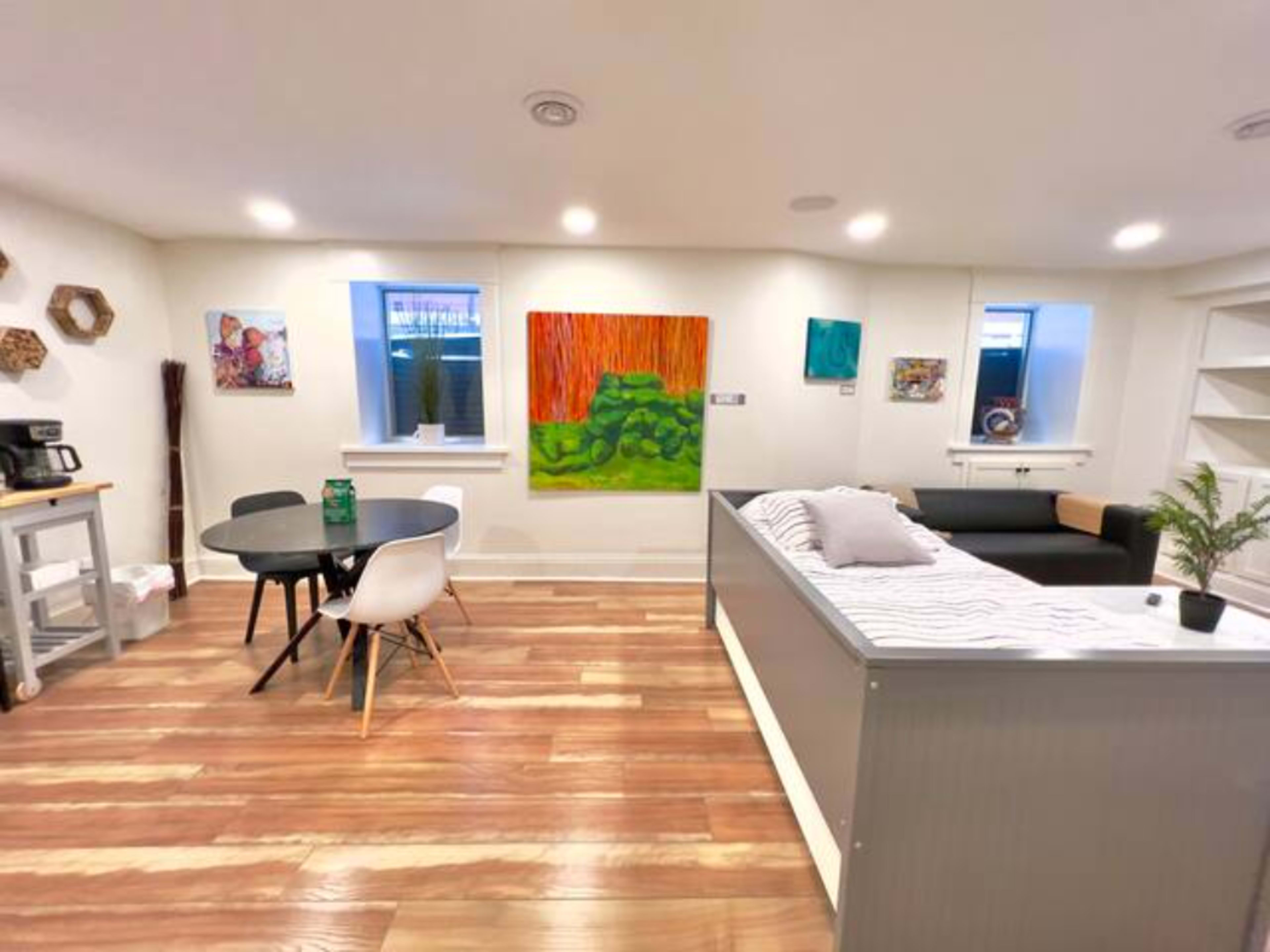 The image shows a modern, minimalistic living space featuring a bed, a small dining table with chairs, and various artworks on the walls.