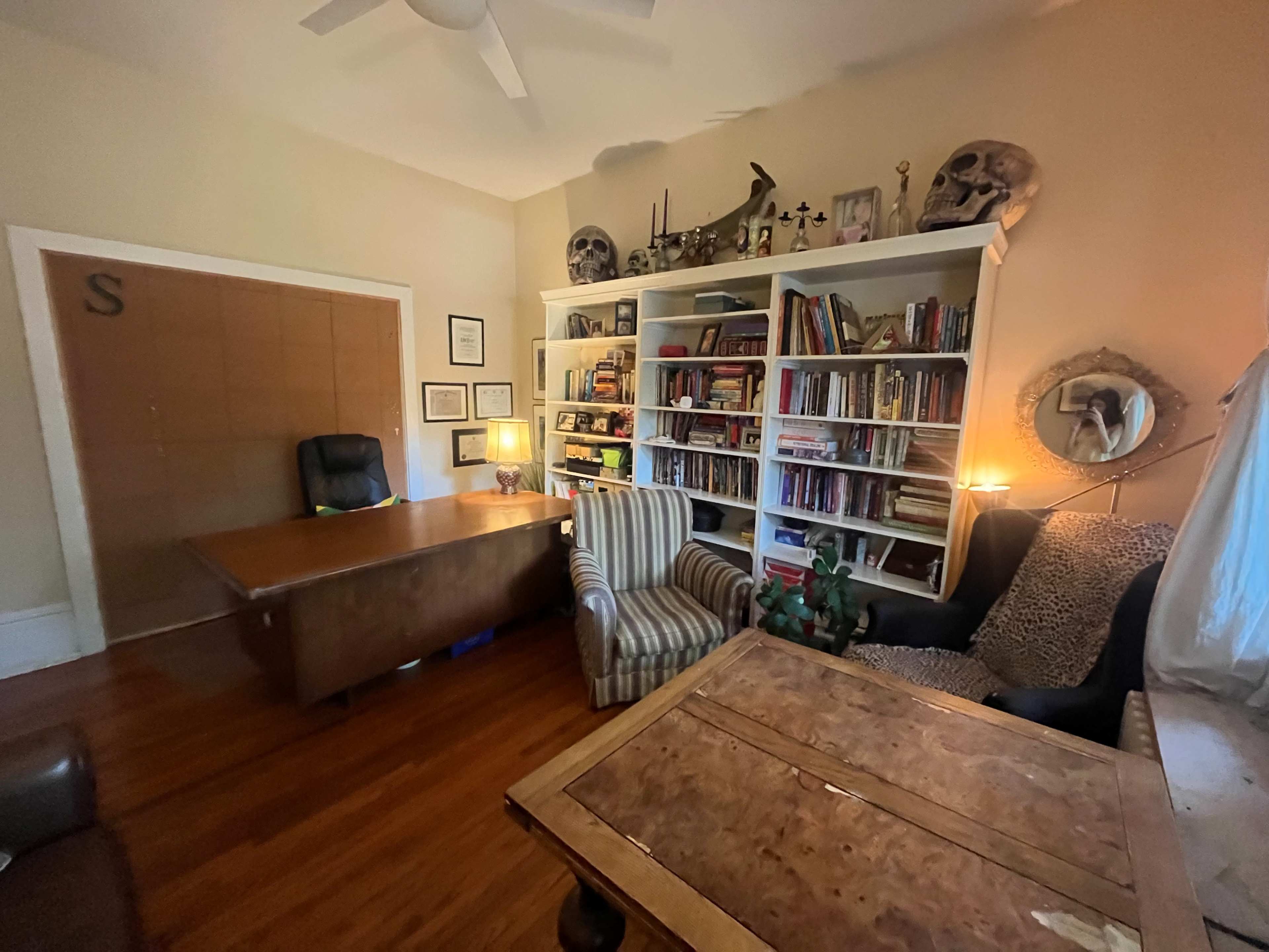 The image depicts a cozy office with a wooden desk, bookshelves filled with books, two armchairs, and a table, all set against a backdrop of light-colored walls.