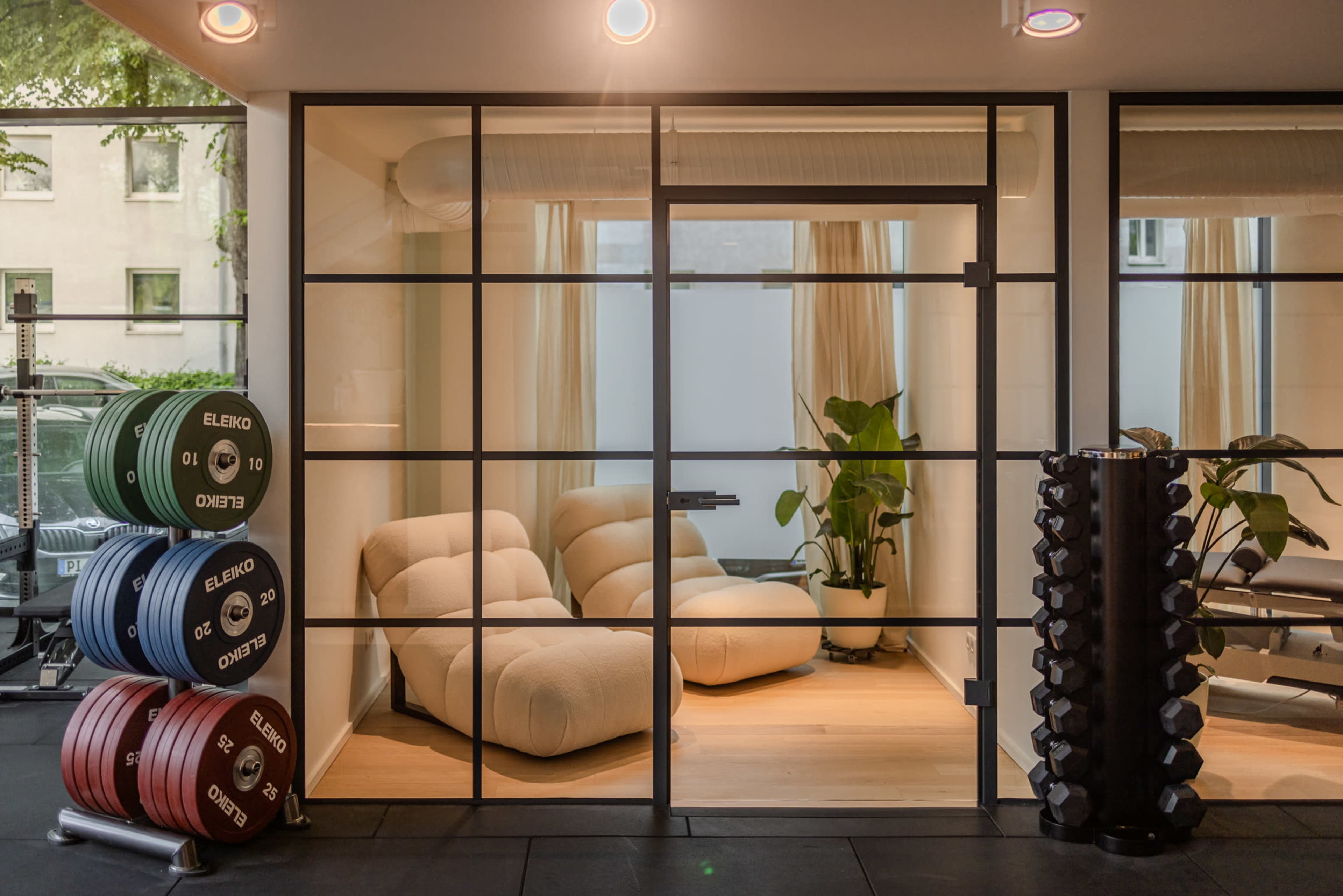 The image shows a modern gym area with a glass-walled relaxation space featuring two beige lounge chairs and greenery, alongside weightlifting equipment stacked against the wall.