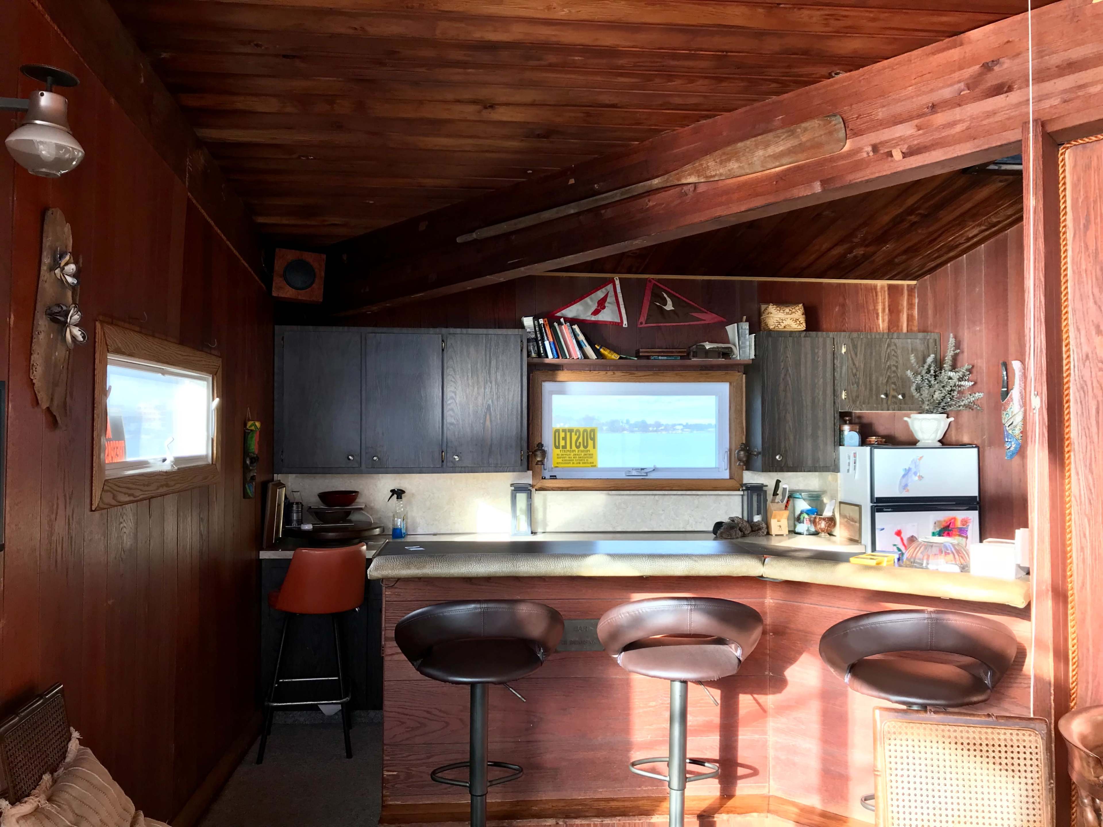 A rustic kitchen with wood-paneled walls, a bar area with three stools, and a window overlooking a water view.