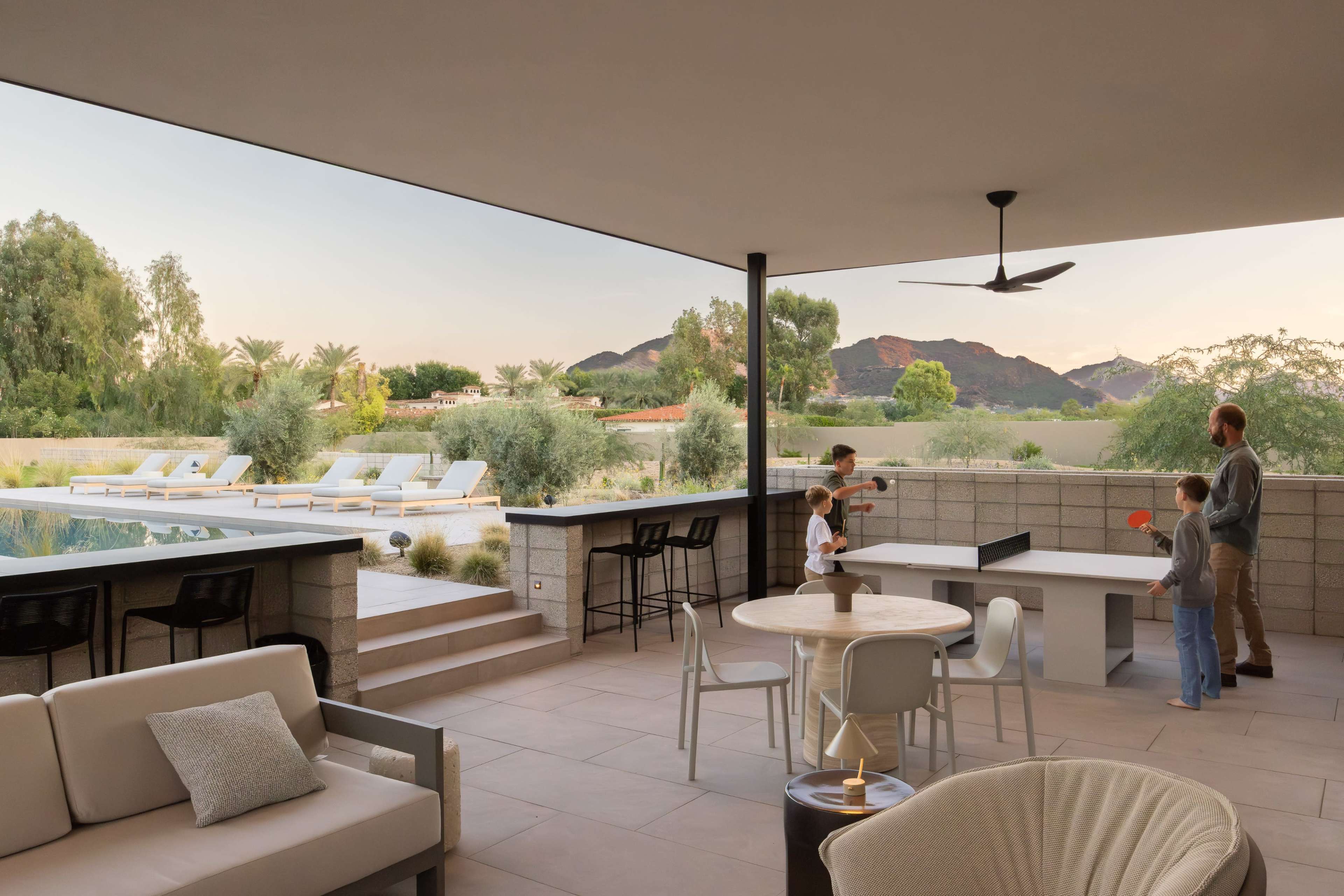 A family engages in a game of table tennis on a covered patio with a view of a landscaped yard and mountains in the background.