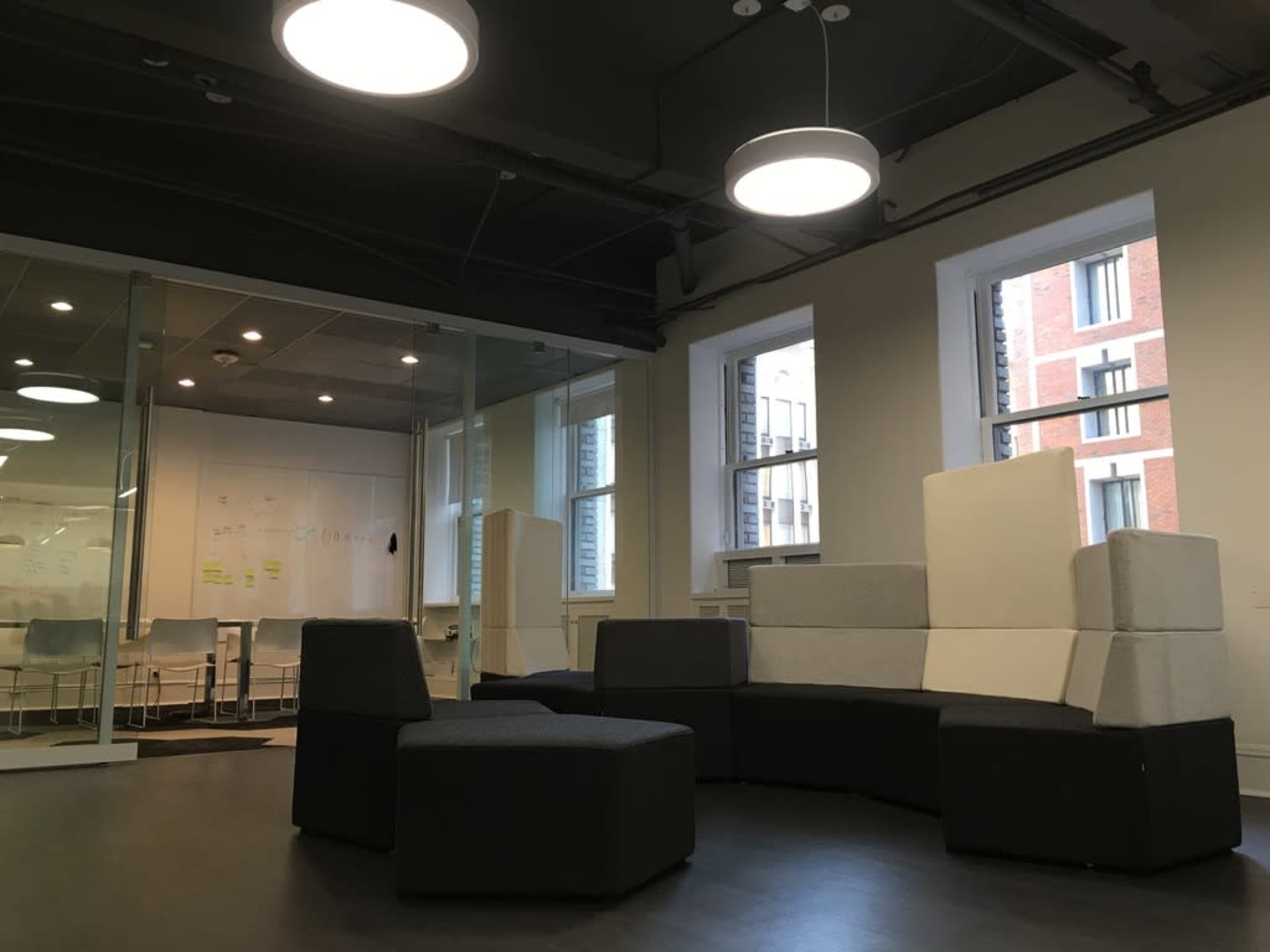 A modern workspace featuring a seating area with modular furniture in gray tones and large windows, along with a glass-walled meeting room in the background.