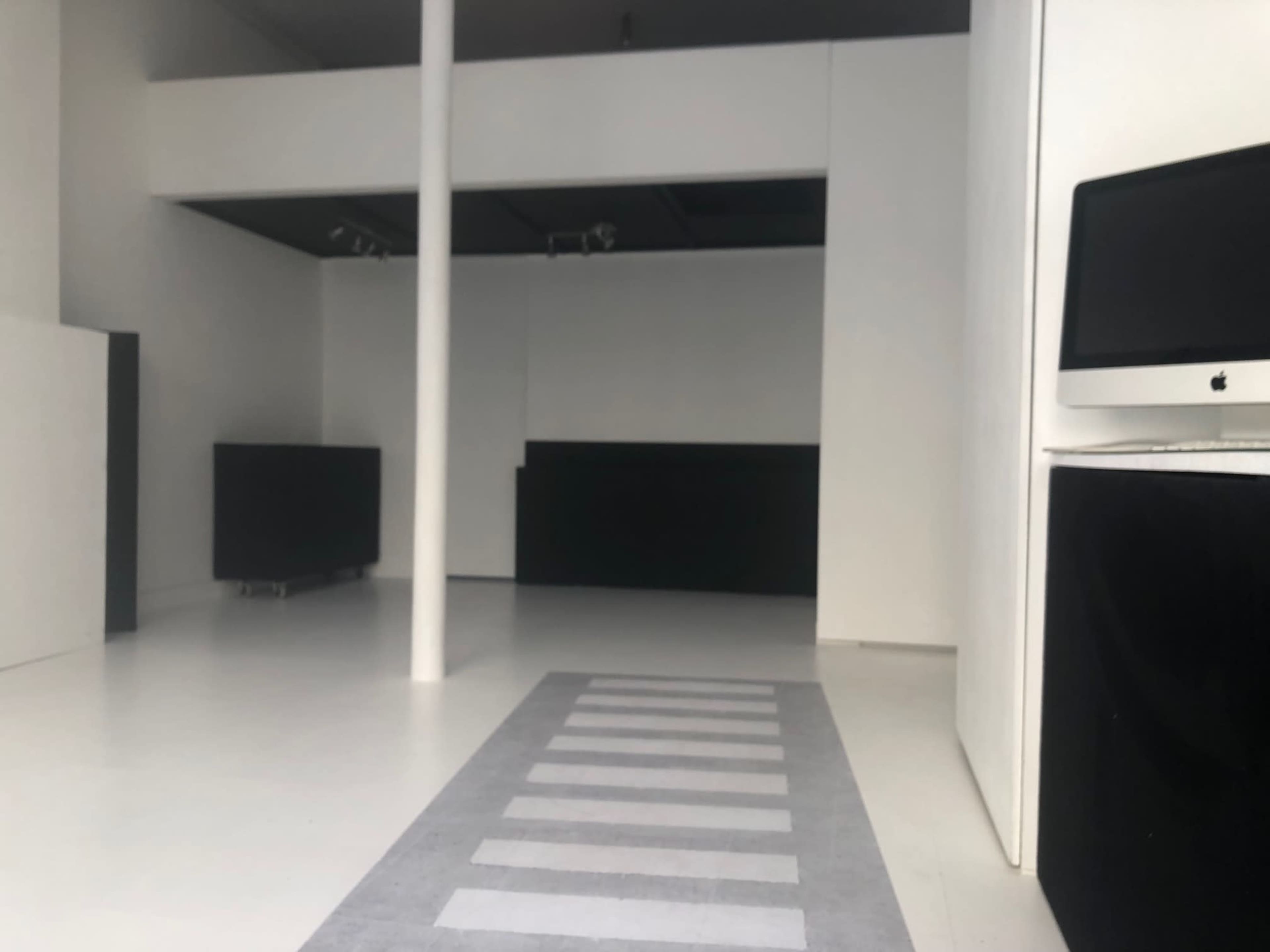A minimalistic, empty interior space featuring white walls, a light floor, and a gray striped rug leading towards a black booth area.