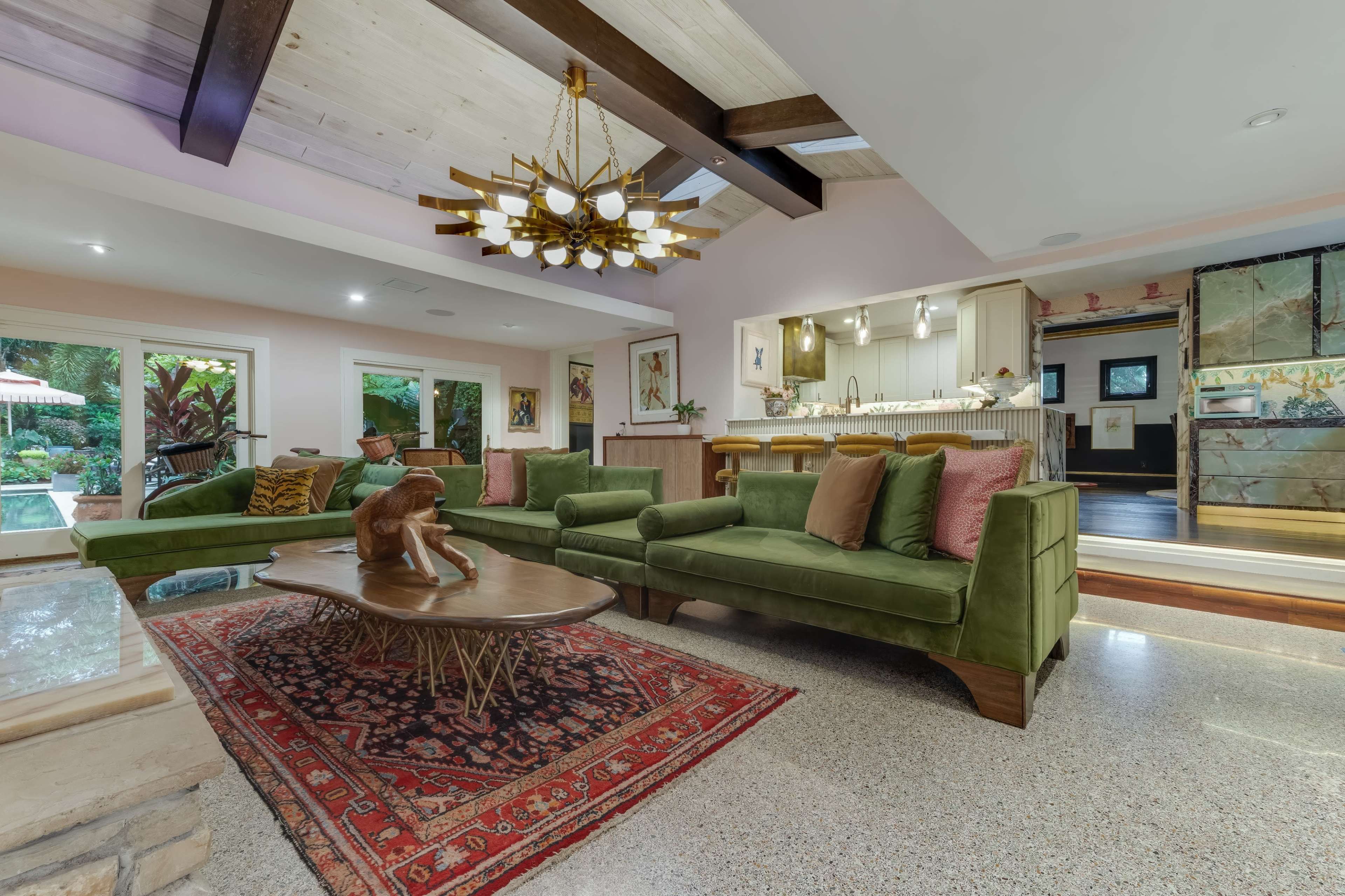 The image shows a modern living room with a green sectional sofa, a wooden coffee table, and a view into an open kitchen area with large windows overlooking a garden.