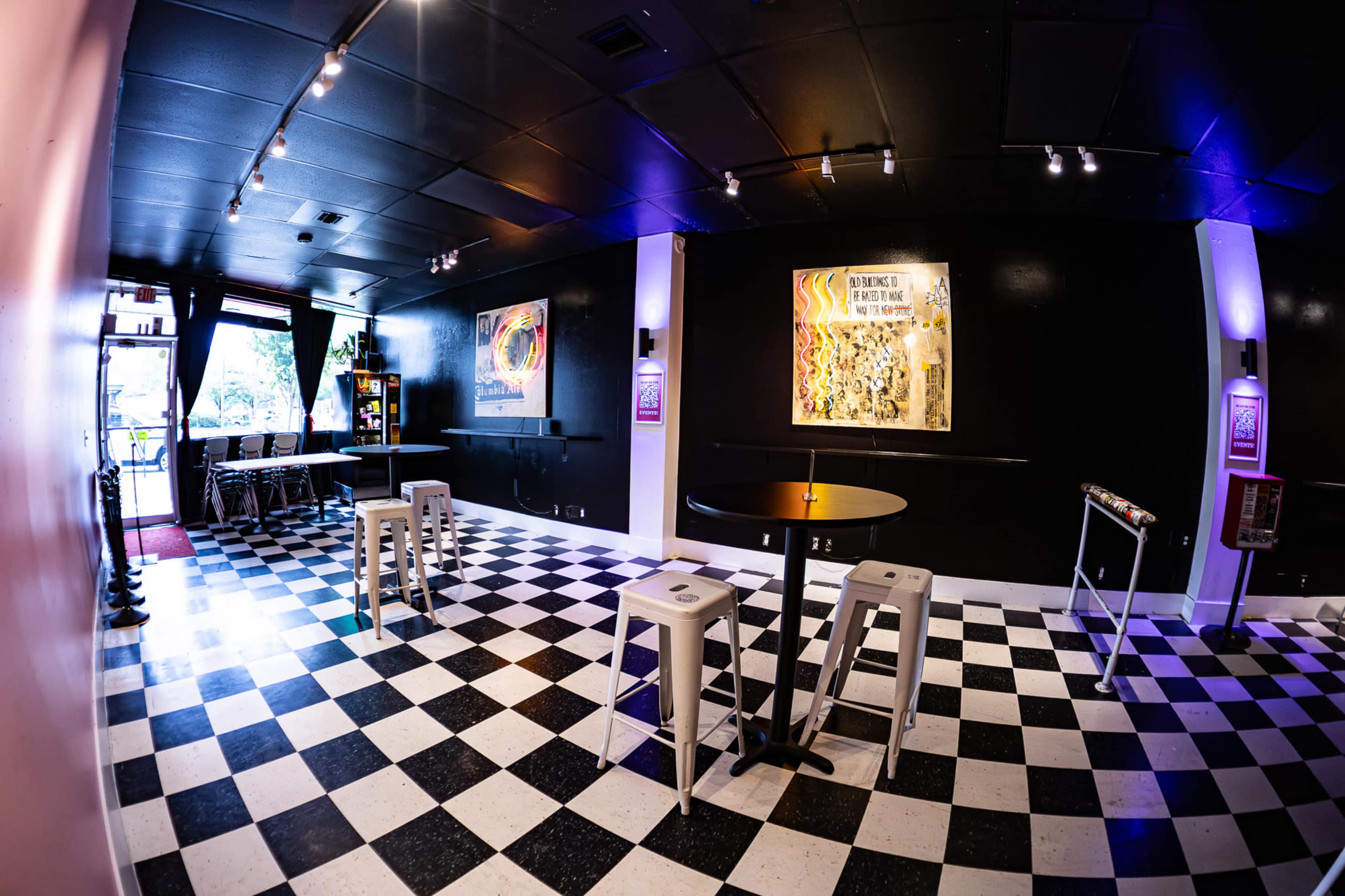 The image shows a modern interior of a café or lounge featuring black and white checkered flooring, high-top tables, and artwork on the walls.