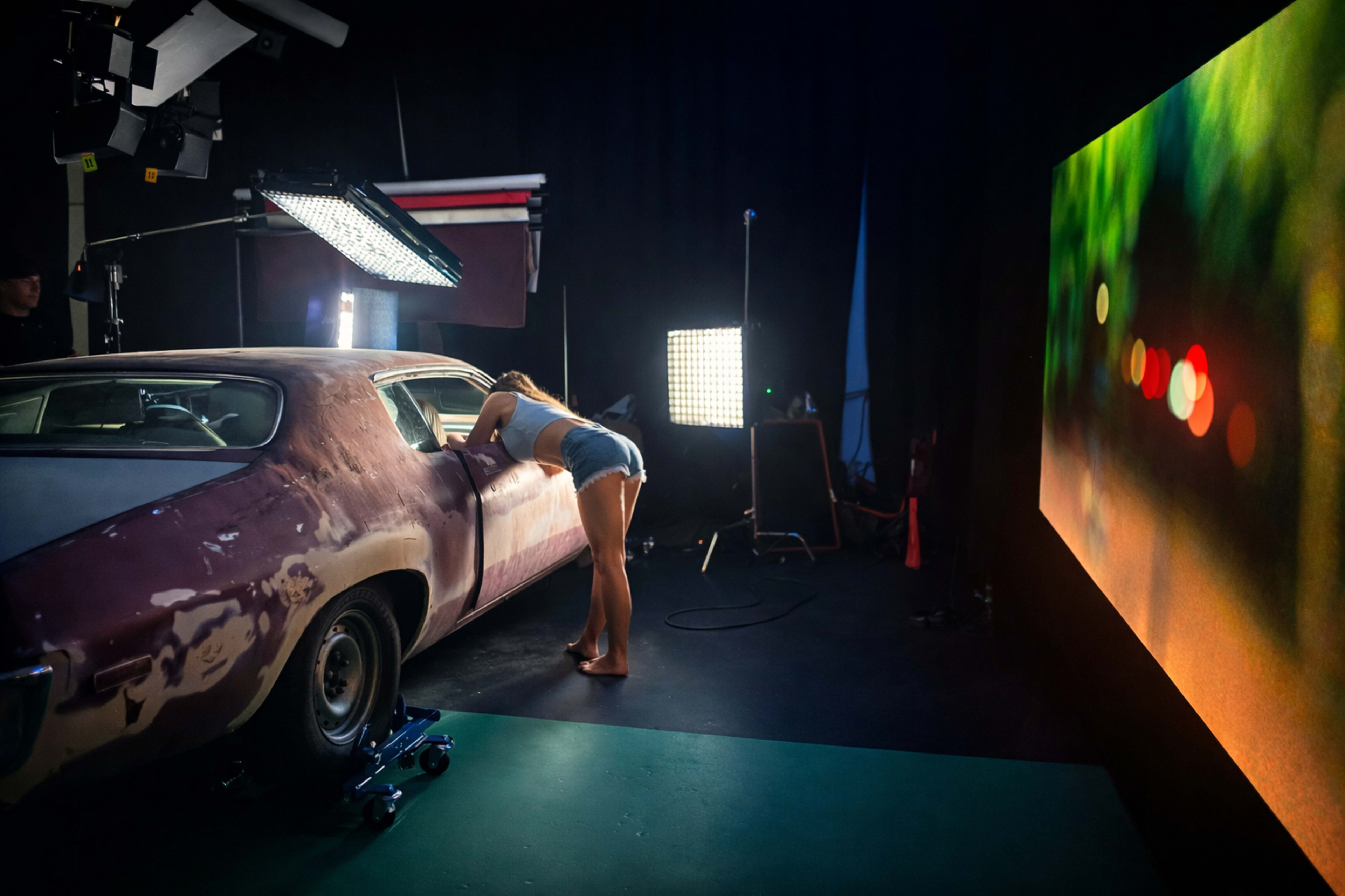 A person leans over a vintage car in a studio setting, surrounded by lighting equipment and a large screen displaying a blurred cityscape.