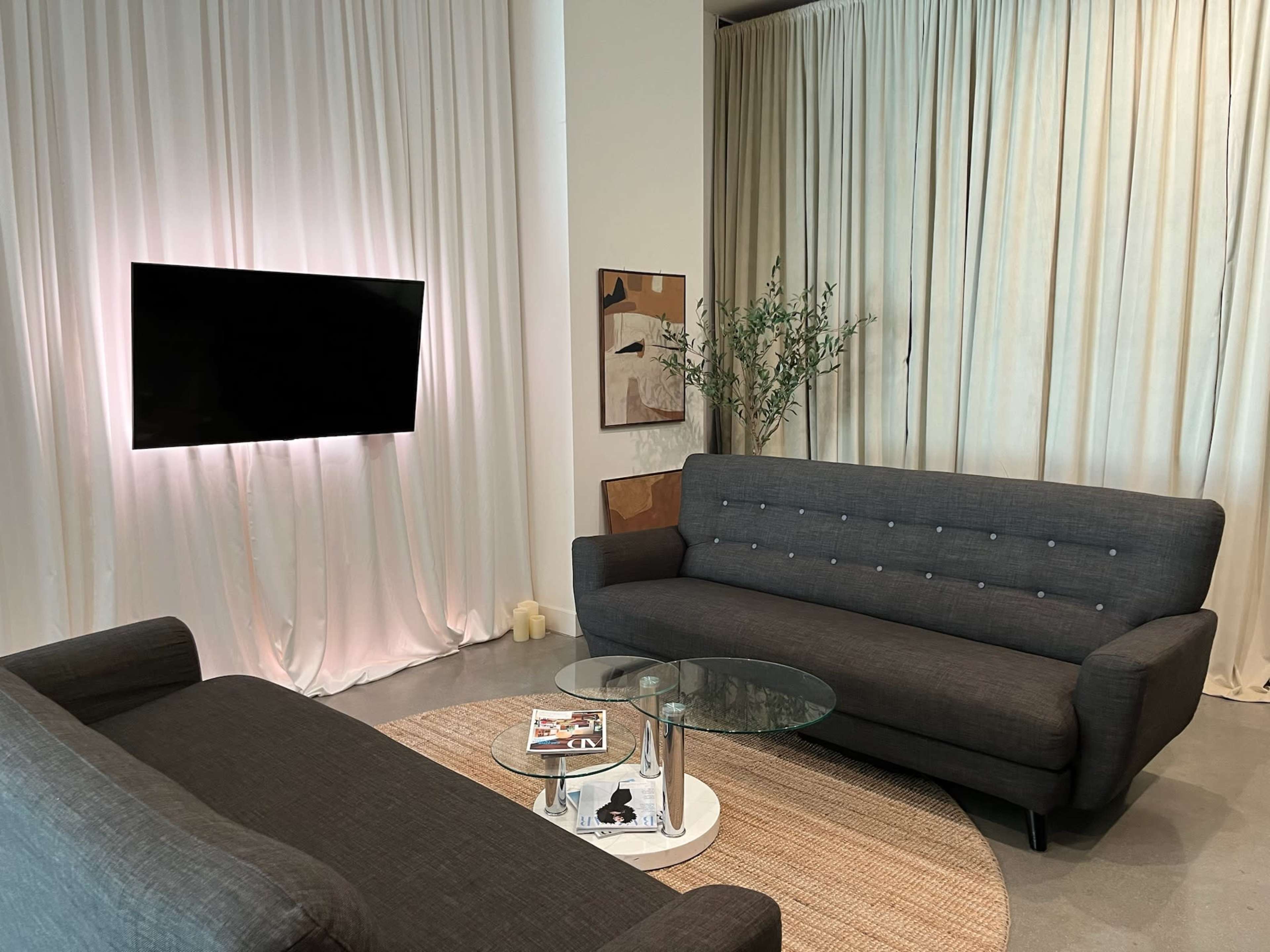 A modern living room features two gray sofas facing a glass coffee table, with a television mounted on the wall and draped curtains in the background.