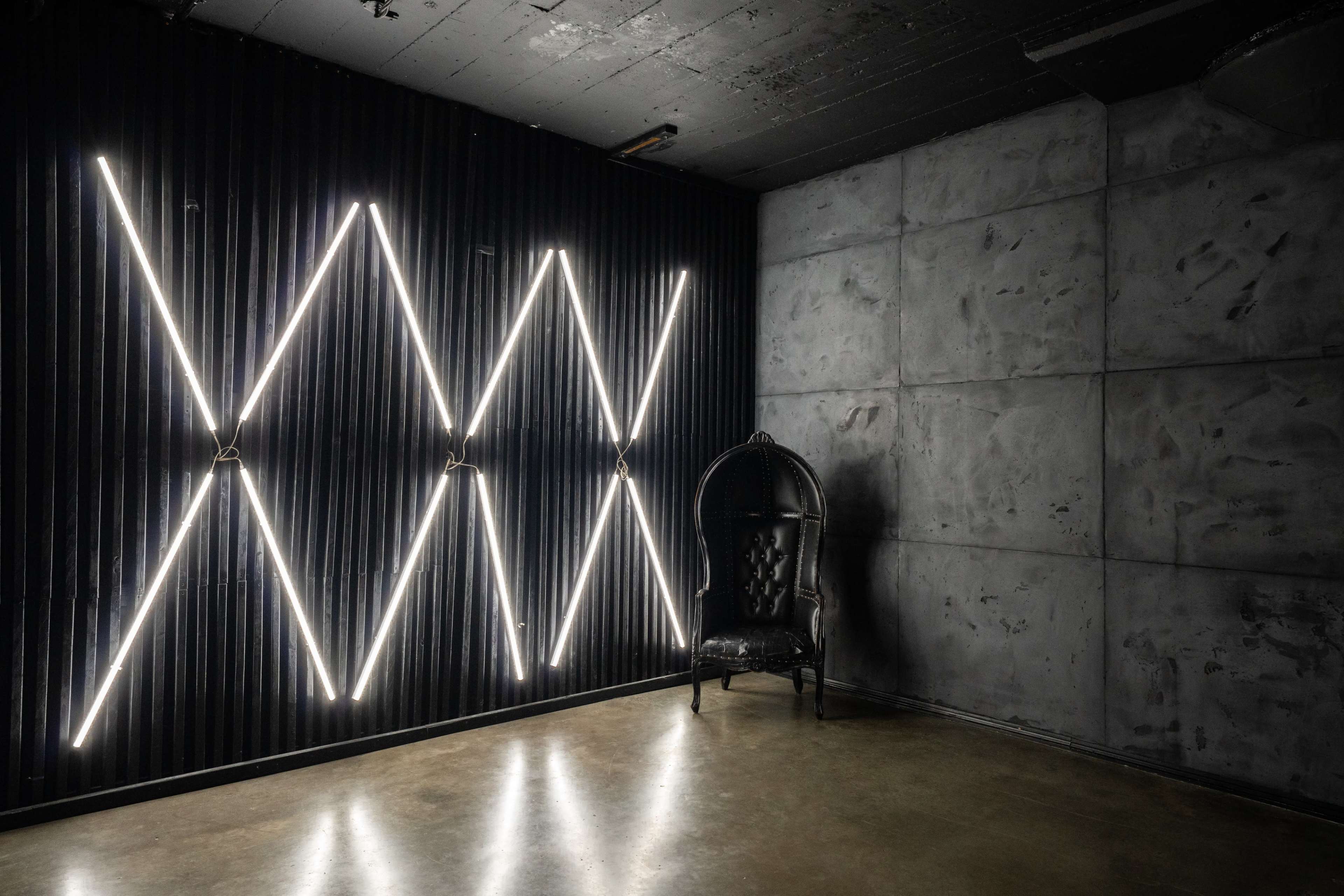 The image shows a minimalist room with a textured concrete wall, a black ornate chair in the corner, and illuminated diagonal light fixtures against a corrugated metal backdrop.