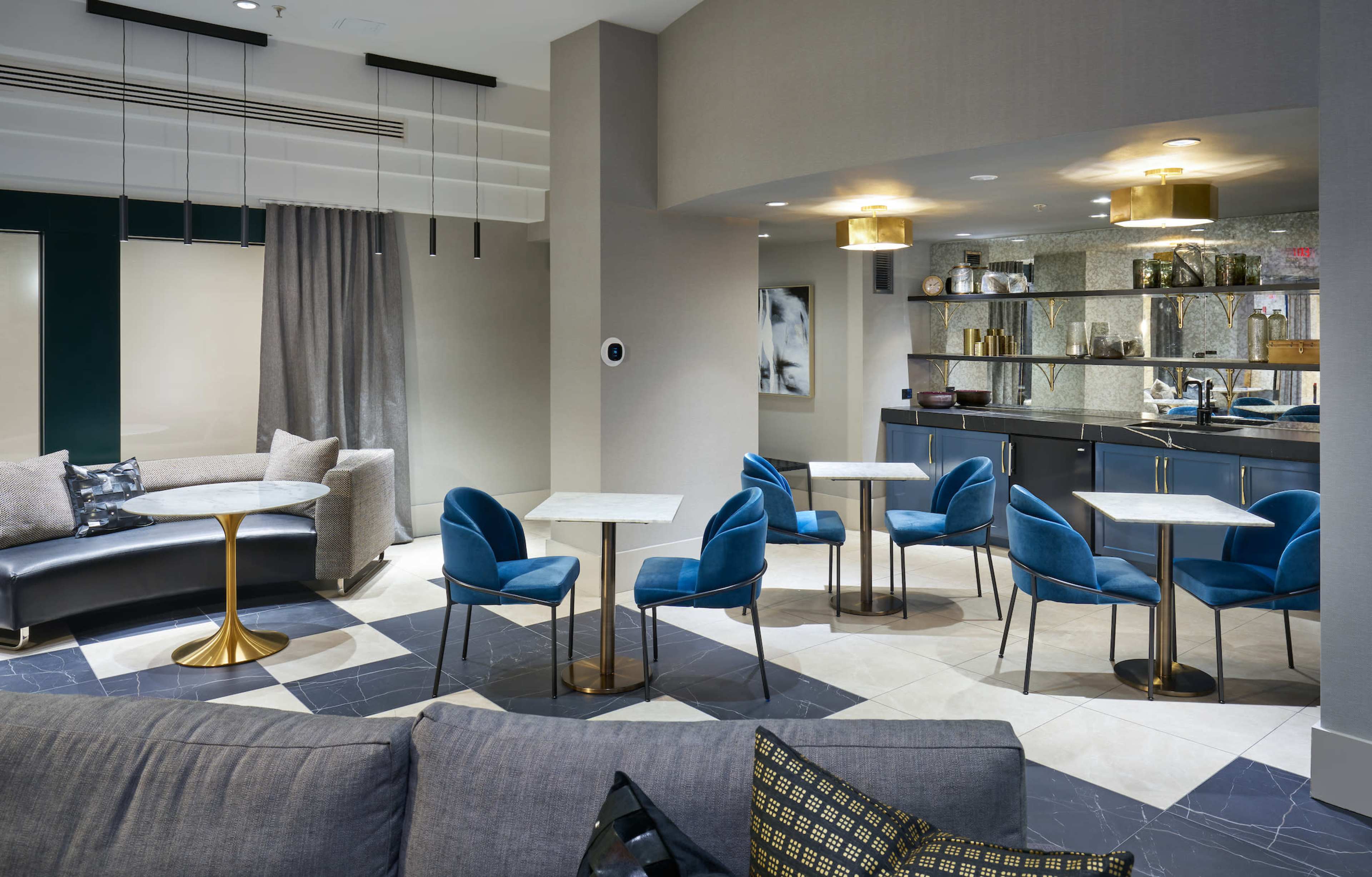 The image shows a modern lounge area featuring blue chairs and marble-topped tables arranged around a stylish space with a gray sofa and a reflective glass shelf filled with decorative items.