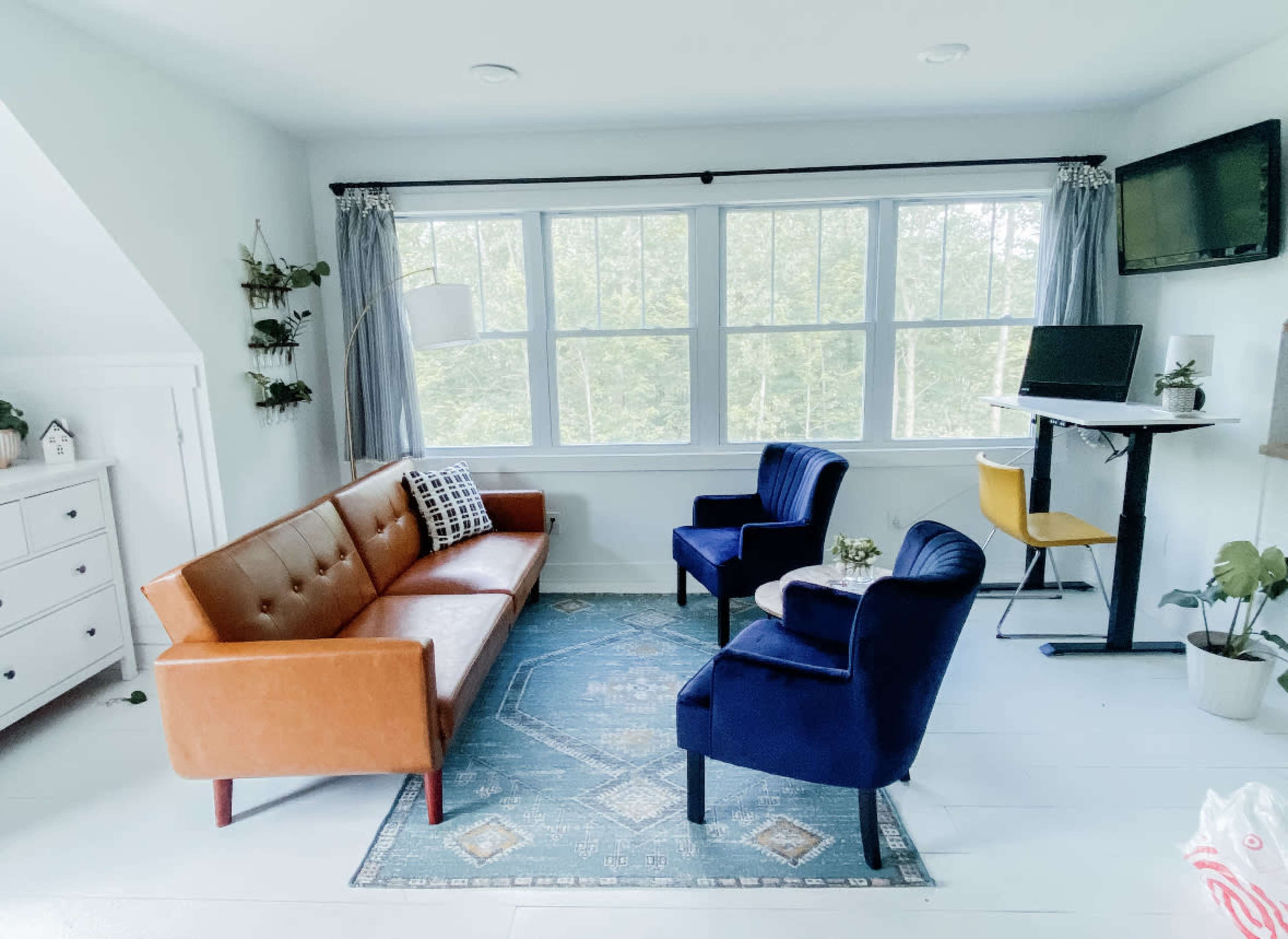 A bright room with a brown leather sofa, two blue chairs, a small table, and large windows that overlook greenery outside.