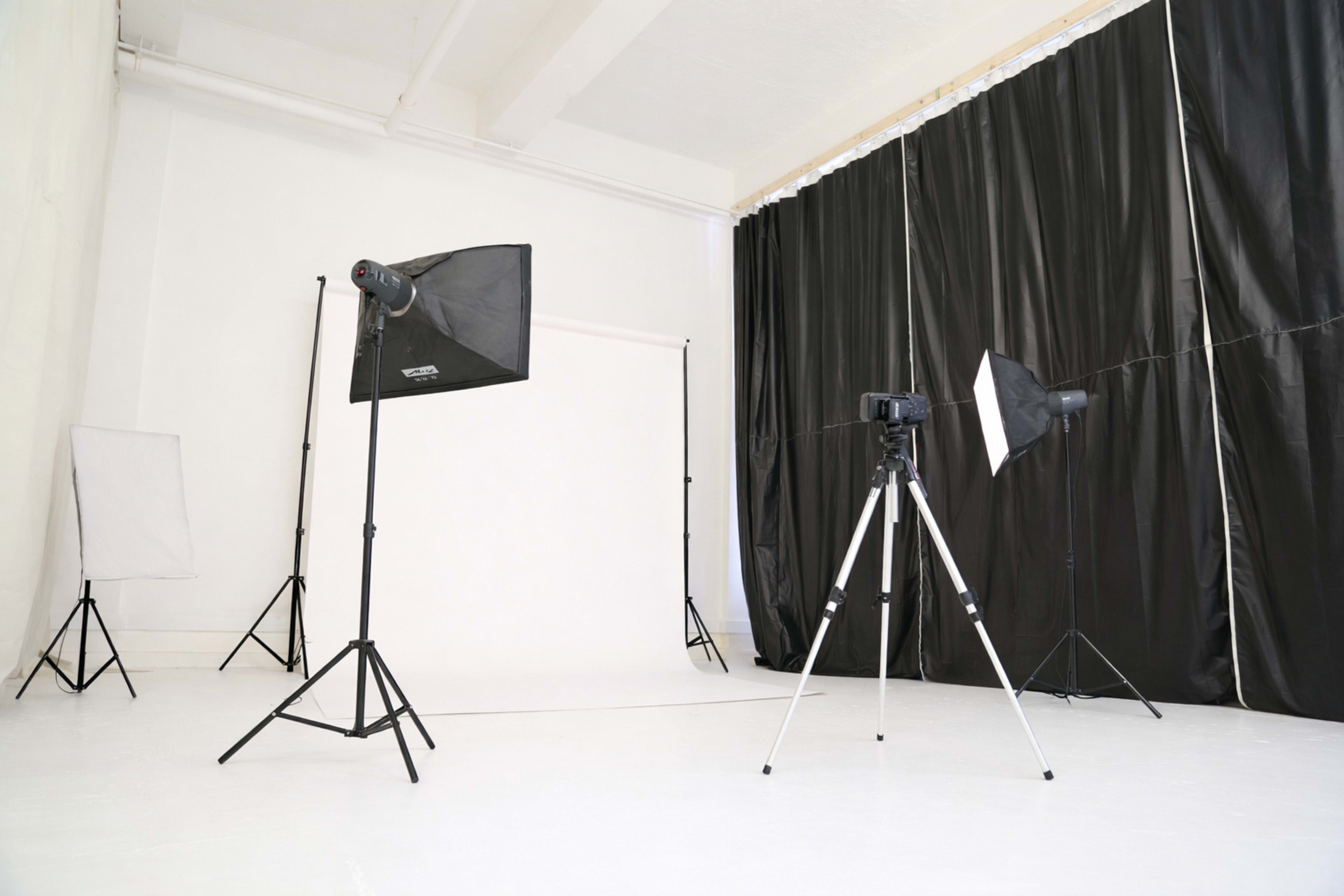The image shows a photography studio with a white backdrop, tripods, and lighting equipment arranged against black curtains.