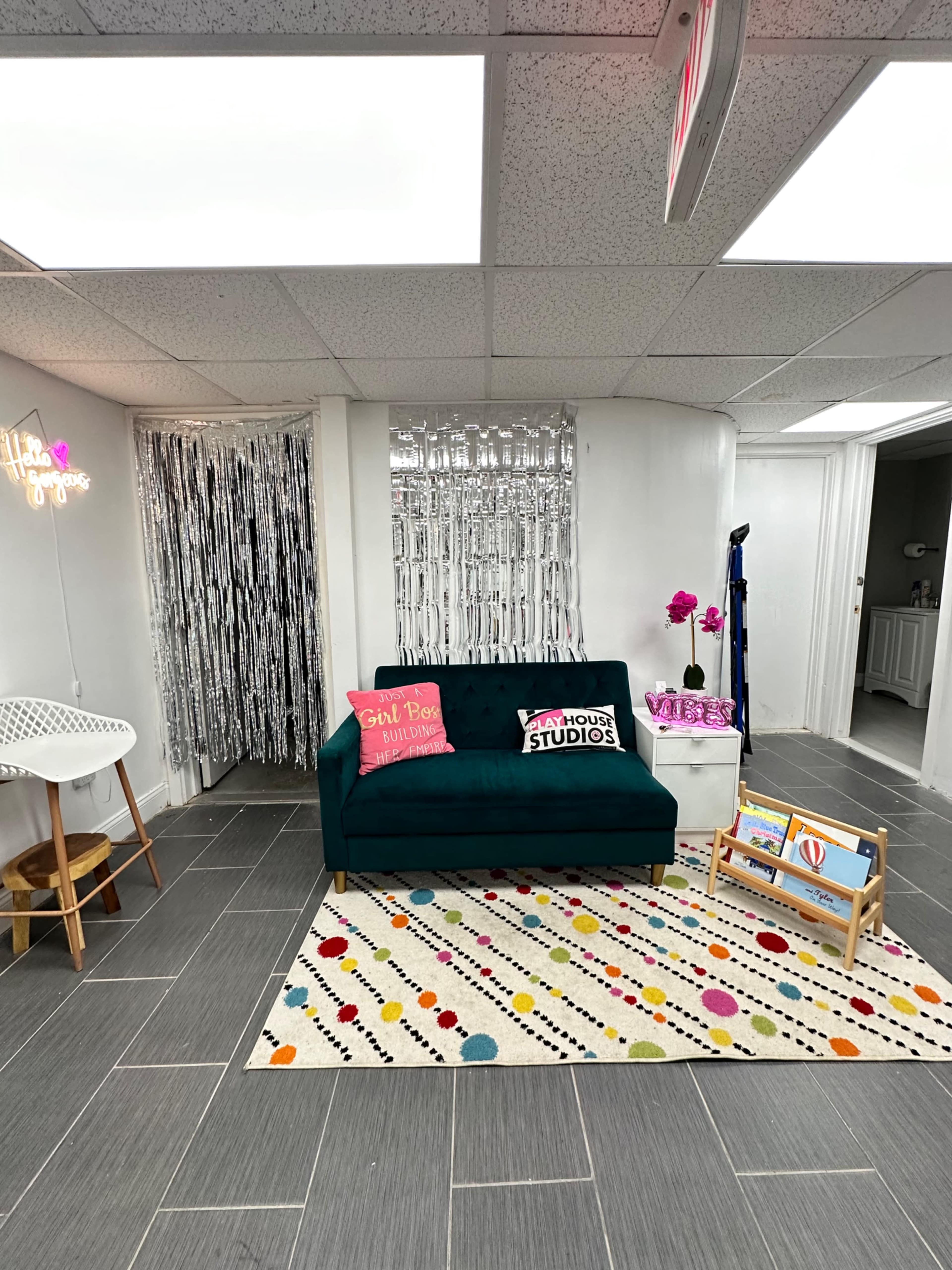 The image shows a small, bright room featuring a teal couch adorned with colorful pillows, a polka-dotted area rug, and a playful art display next to a set of metallic curtains.