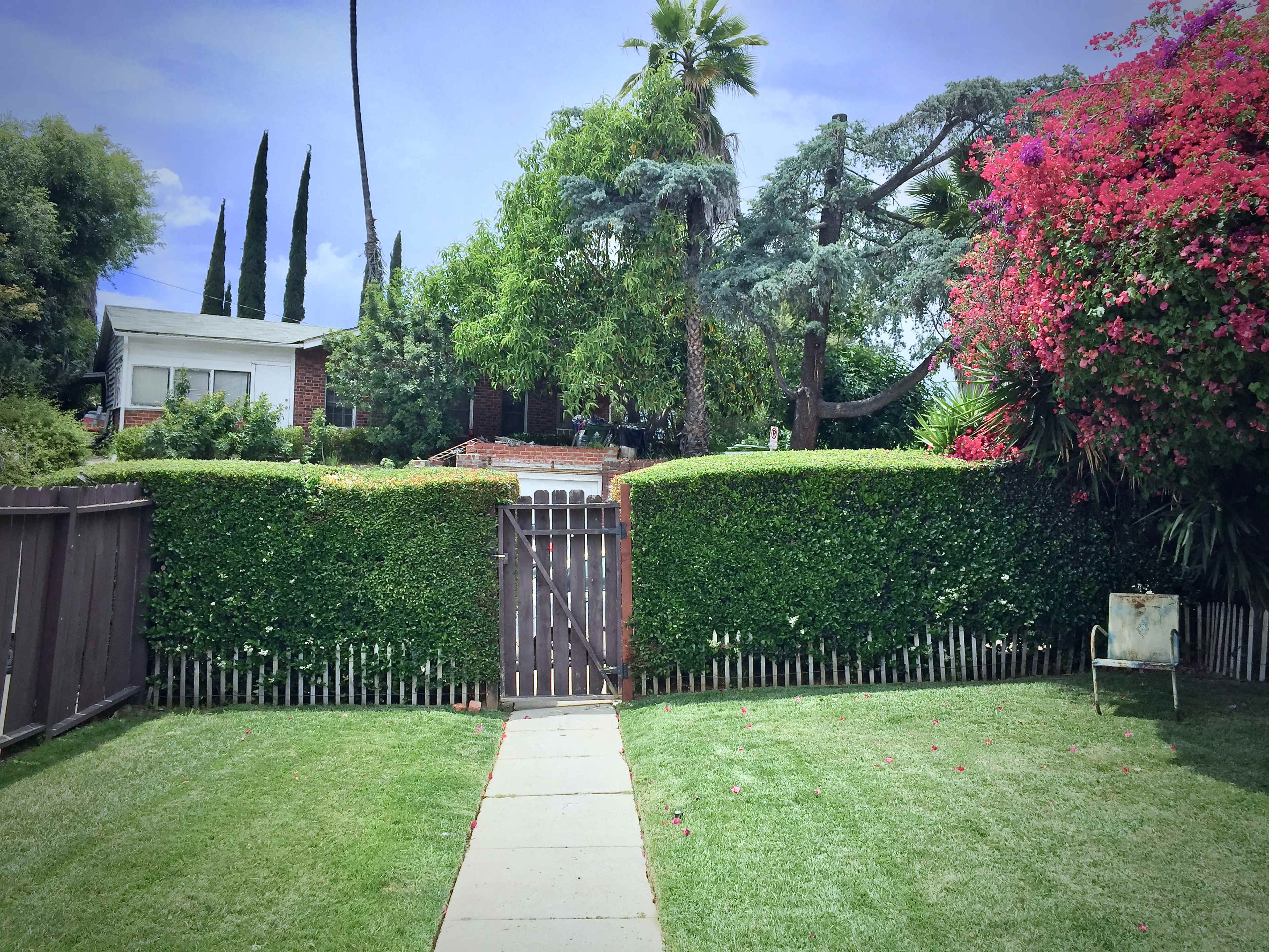 A wooden gate opens onto a neatly maintained lawn bordered by hedges and colorful flowering plants, with a house partially visible in the background.