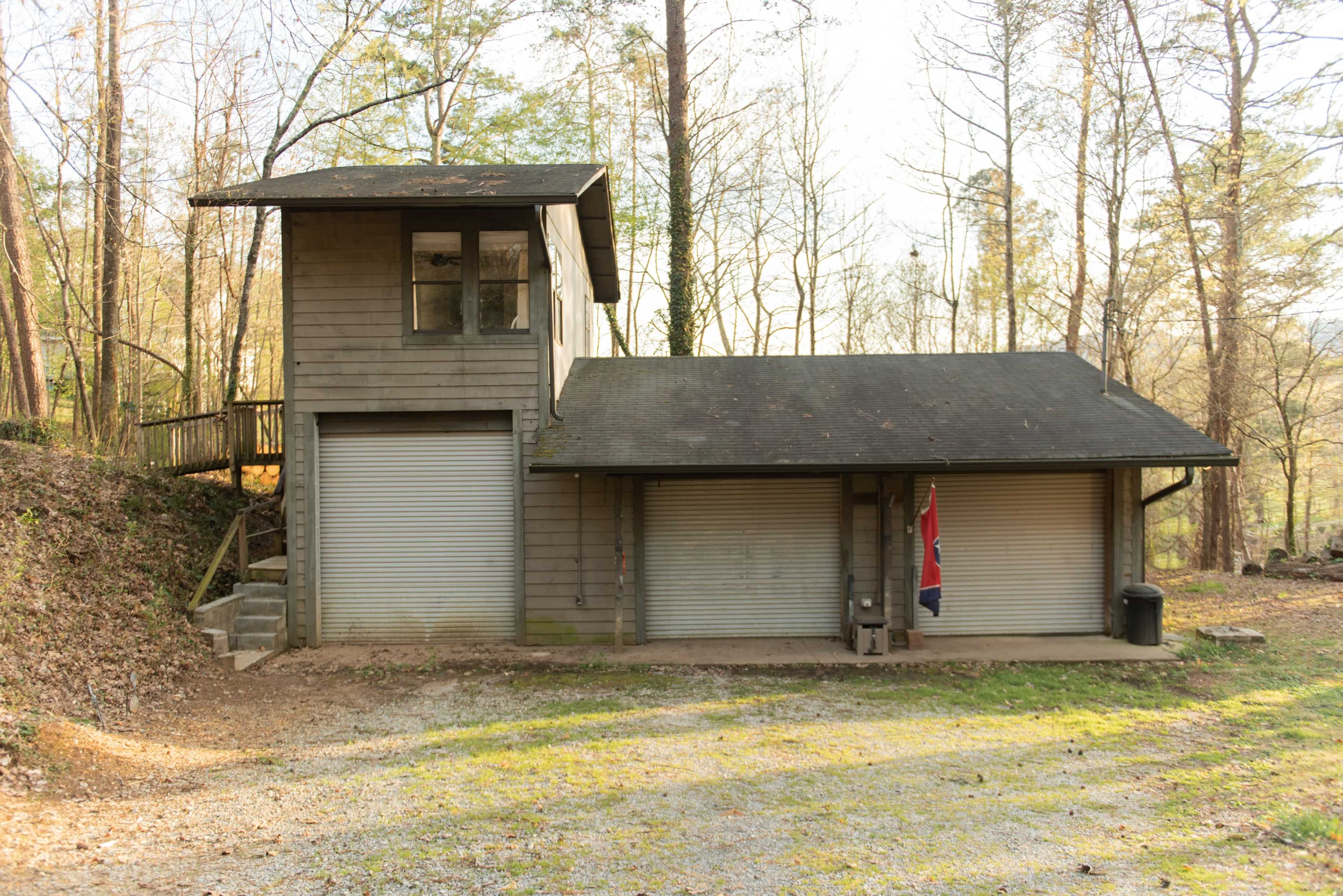 A two-story wooden structure with garage doors, located in a wooded area, featuring a Confederate flag hanging outside.