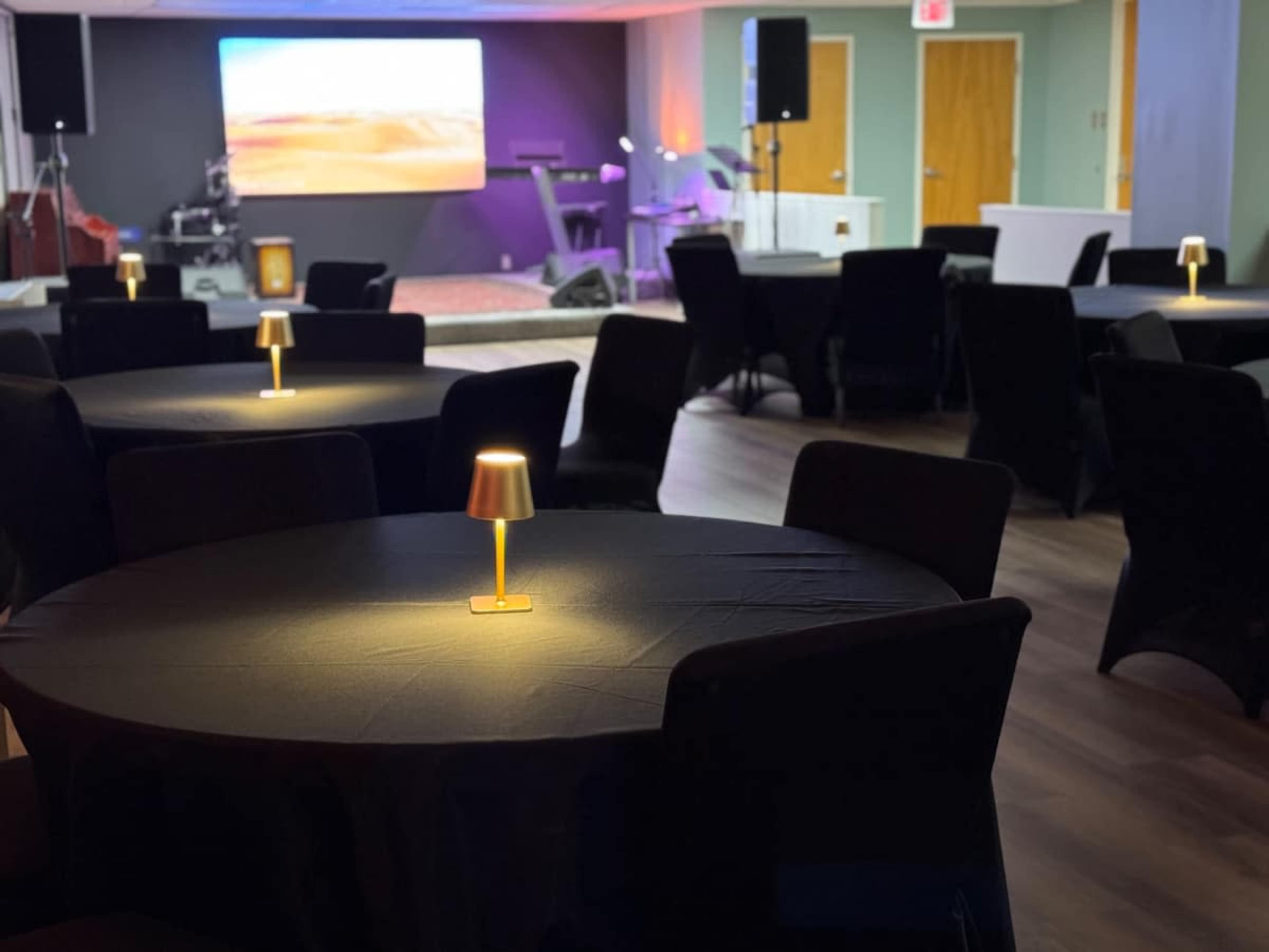 The image shows a spacious event room with round black tables, each featuring a small lamp, and a screen displaying a desert landscape in the background.