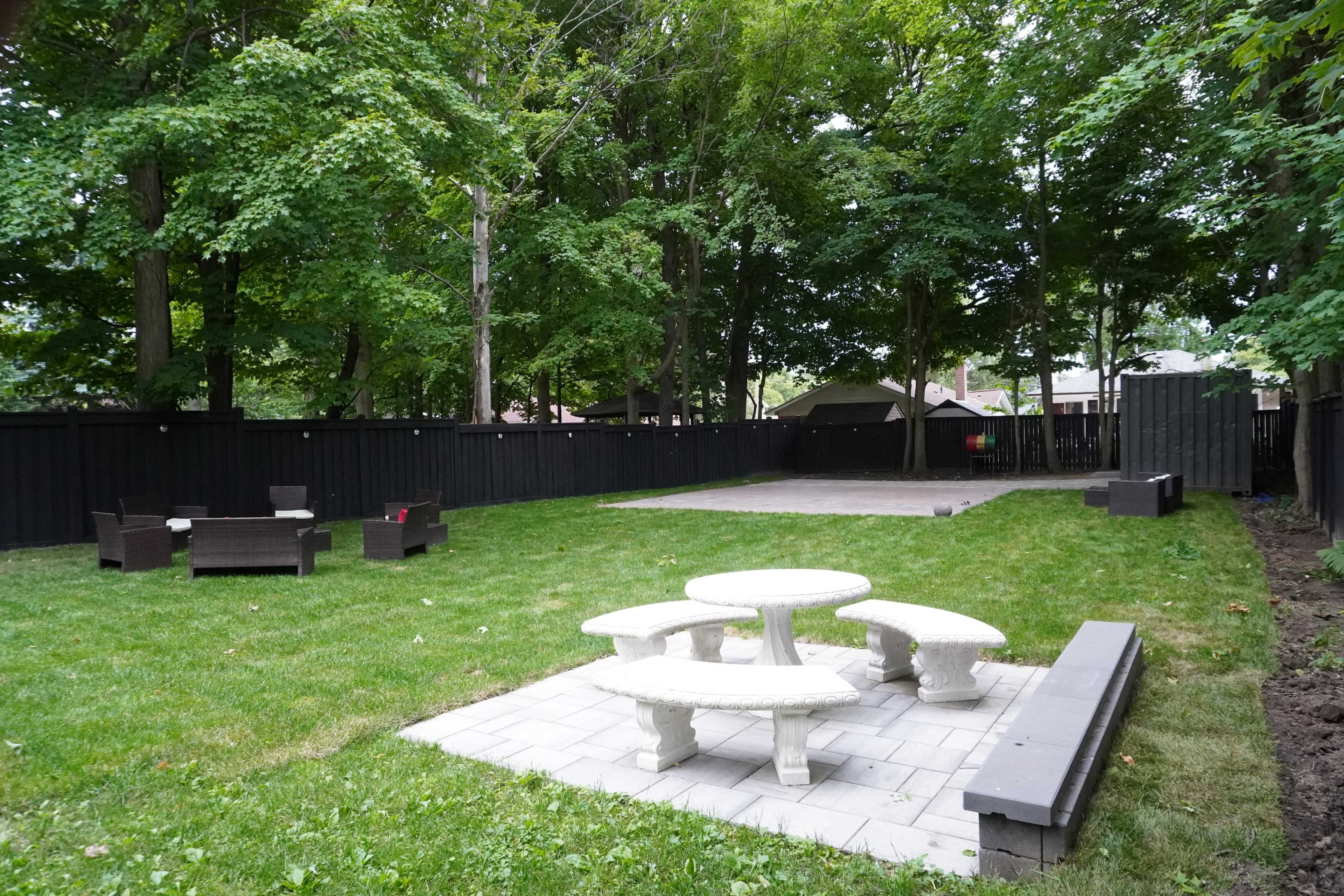 The image shows a spacious outdoor area with a stone table and seating arrangement, surrounded by a green lawn and wooded backdrop.