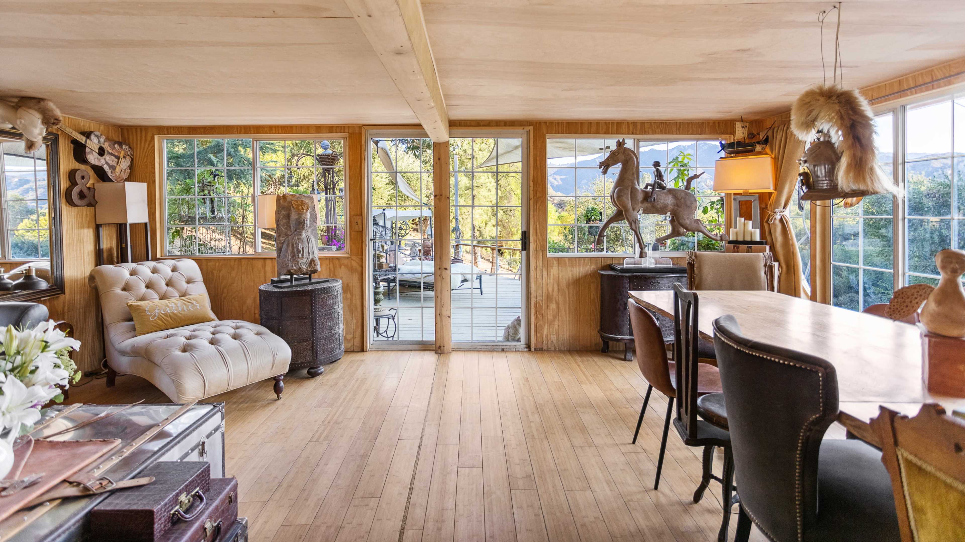 The interior of a spacious, wood-paneled room features a mix of sculptures, a seated area with a tufted chair, and a dining table near large windows overlooking an outdoor deck.