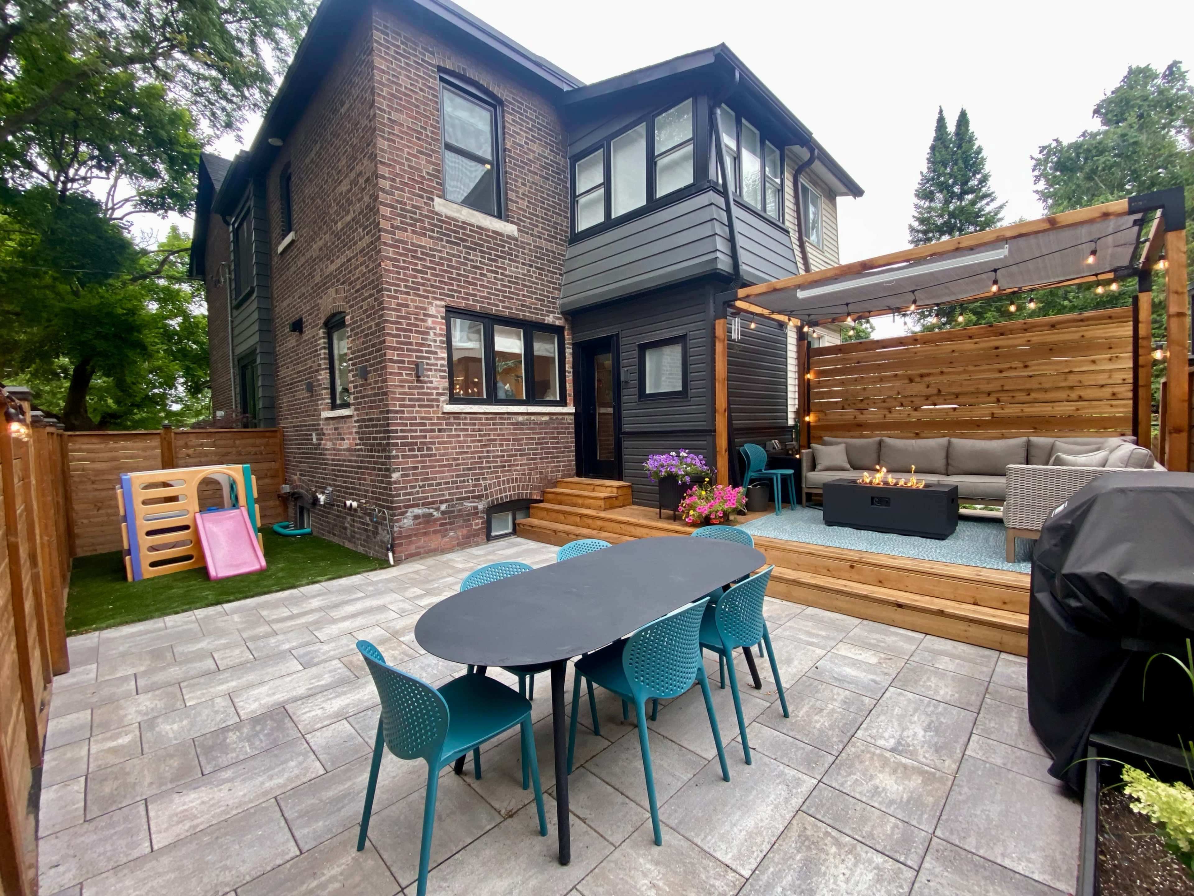 The image shows a backyard patio area featuring a wooden seating arrangement, a dining table with teal chairs, and a brick and wooden house in the background.