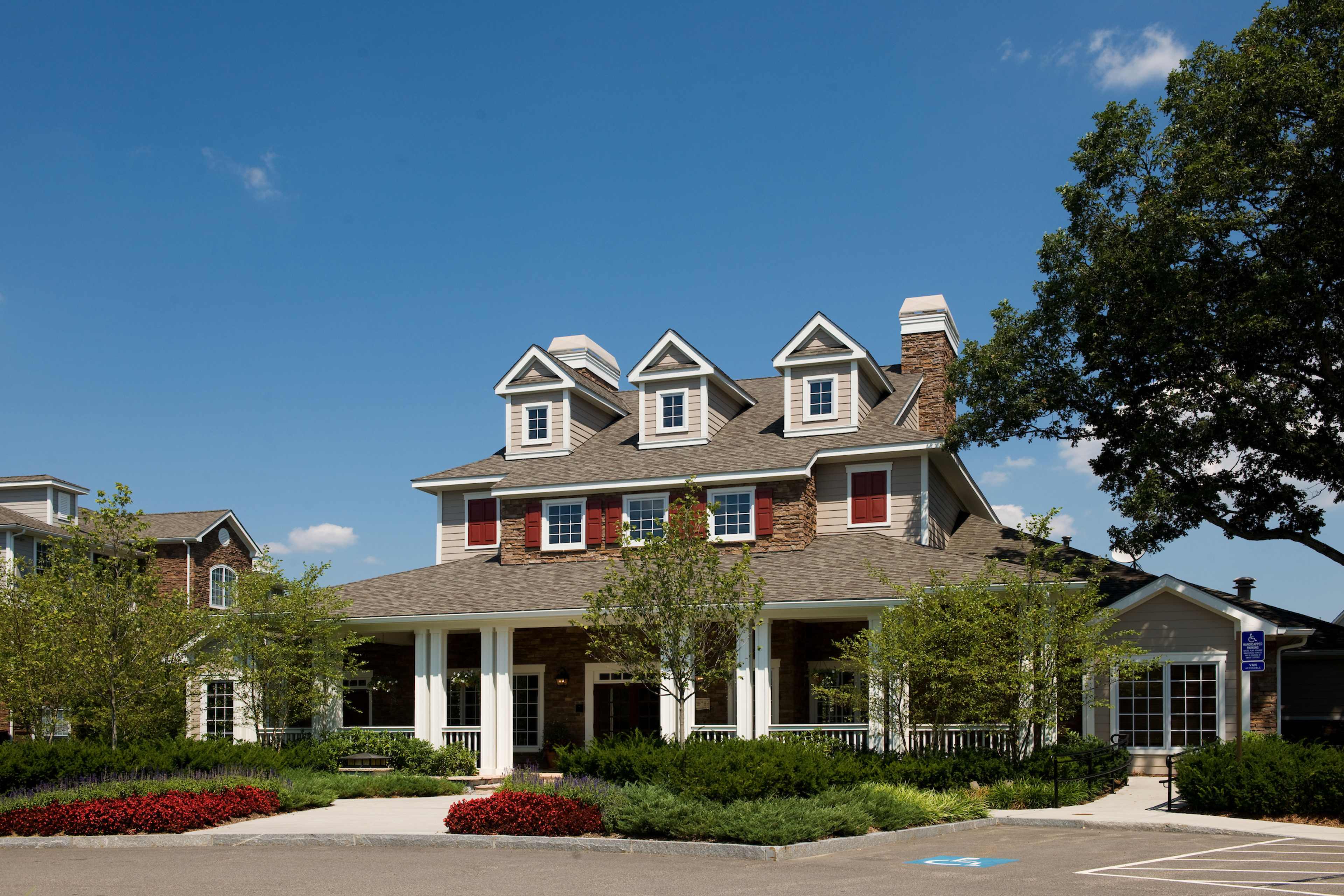 A large, multi-storied house with a stone exterior, gabled roof, and red window shutters is surrounded by neatly trimmed shrubs and trees.