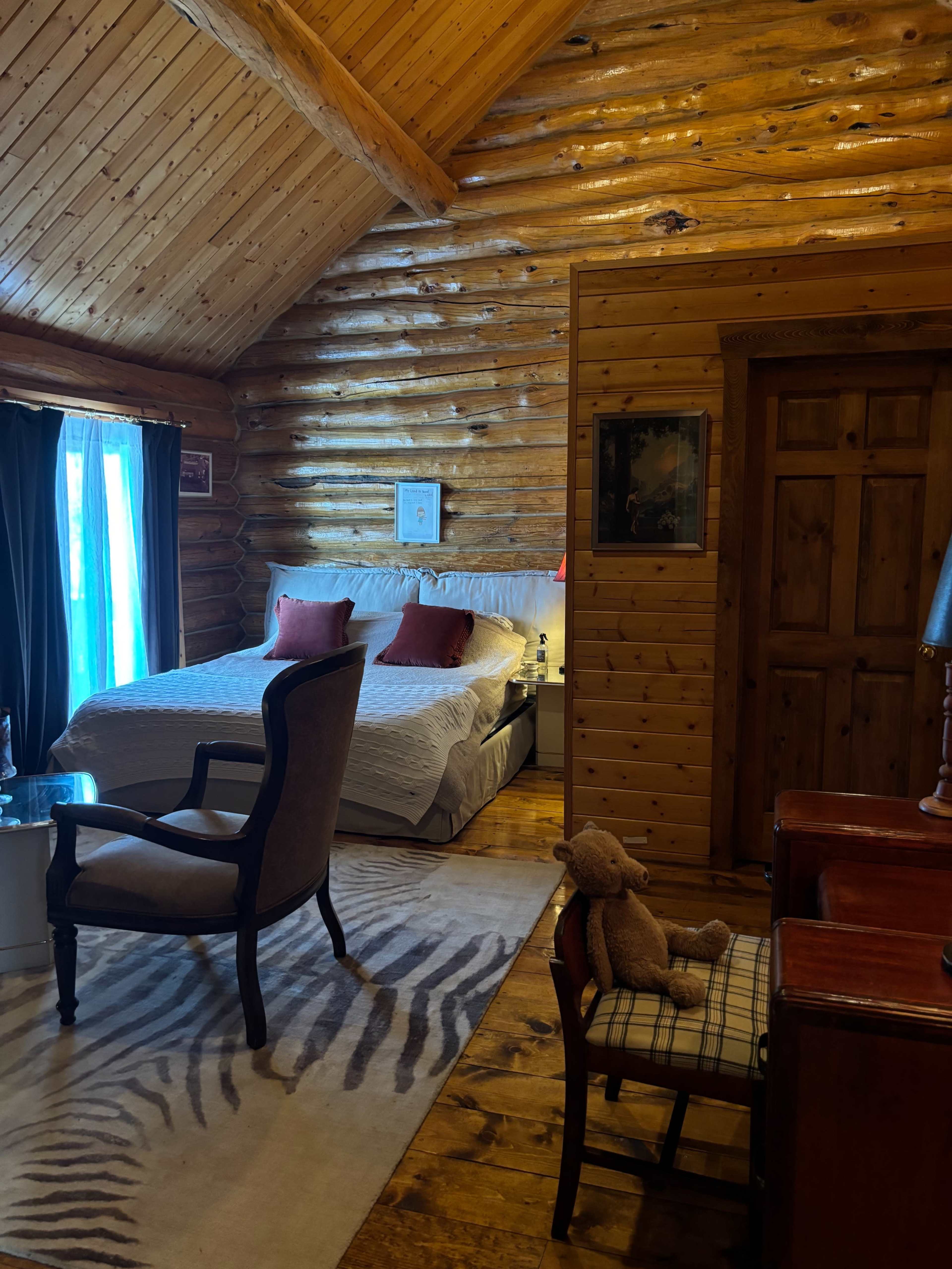The image shows a cozy bedroom with a log cabin design featuring a bed, an armchair, a rustic desk, and a plush bear sitting on a chair.
