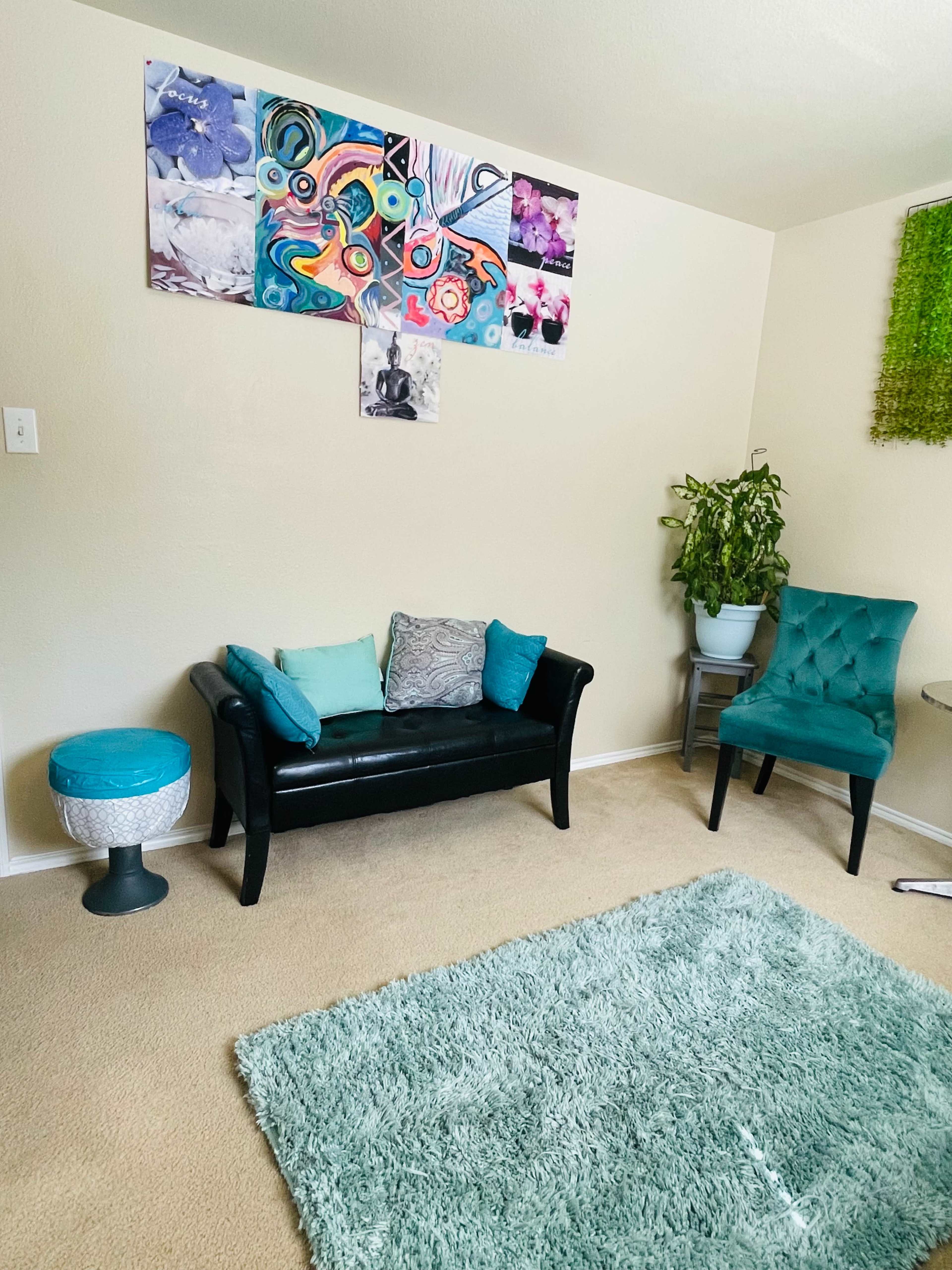 The image shows a small living area decorated with a colorful wall art, a black sofa with teal cushions, a green chair, a round table, and a potted plant.