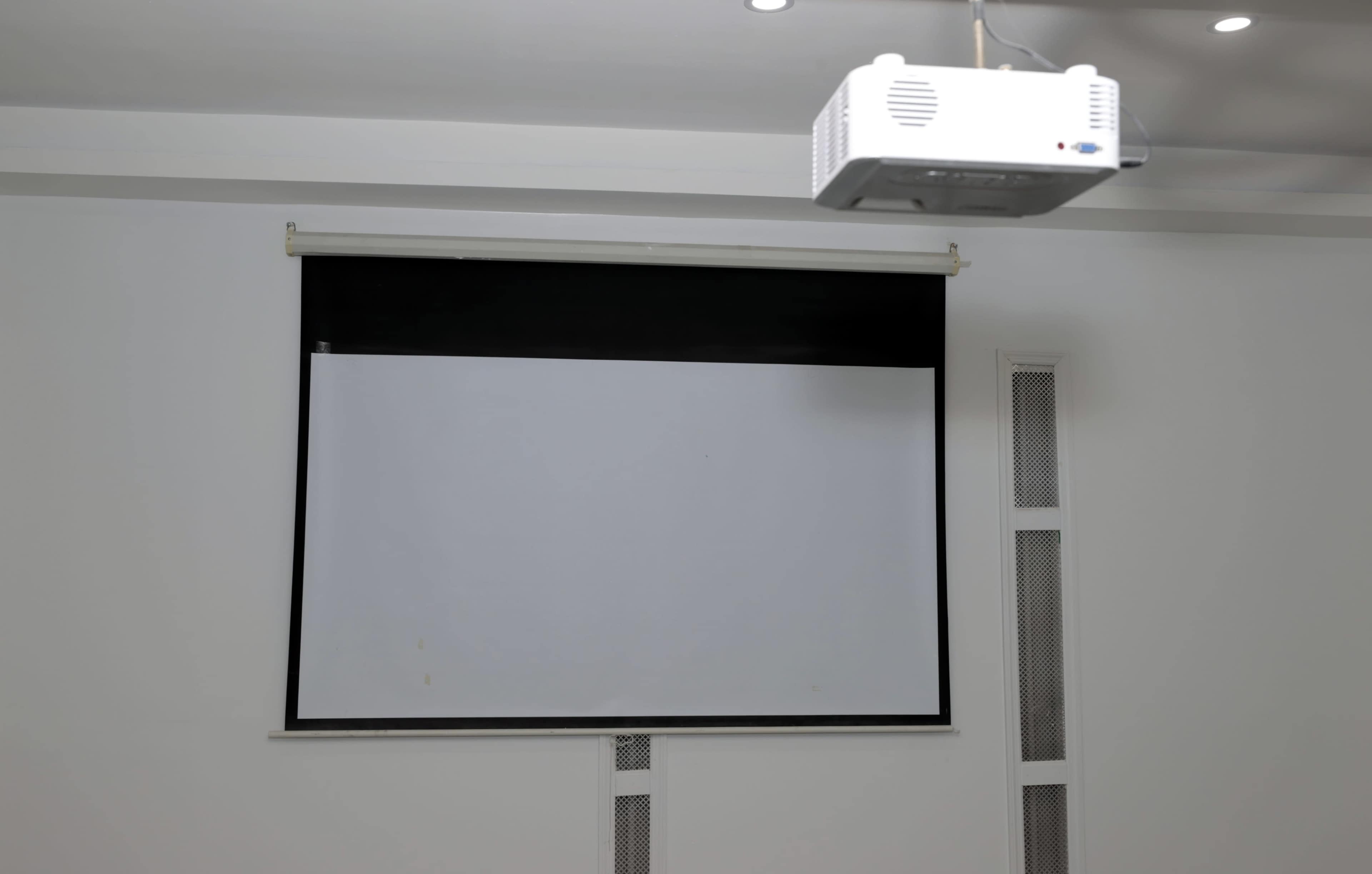 The image shows a blank projection screen mounted on a wall with a ceiling-mounted projector above it.