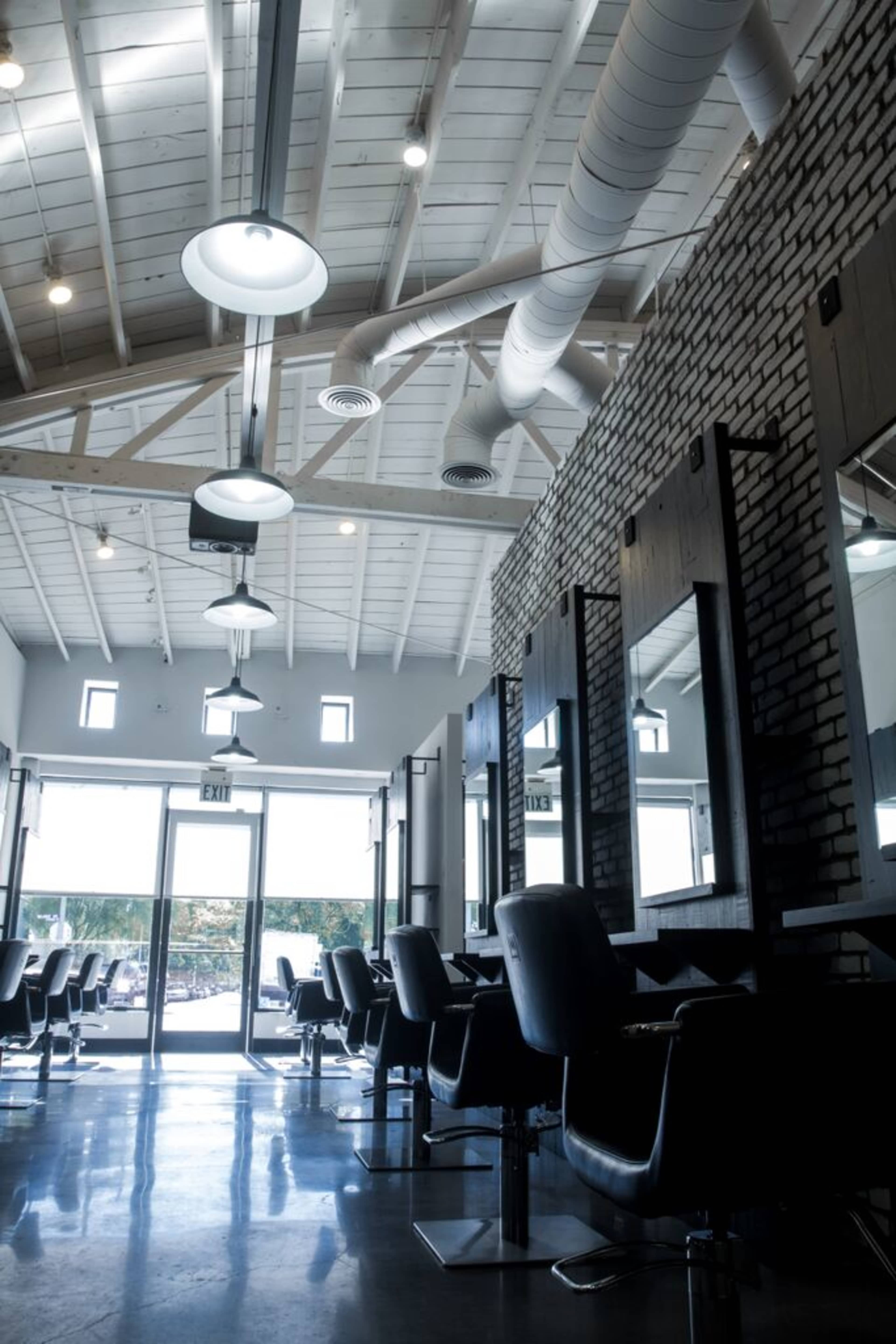 The image shows the interior of a modern salon featuring styling stations, large mirrors, and a bright, open layout with large windows and a high ceiling.